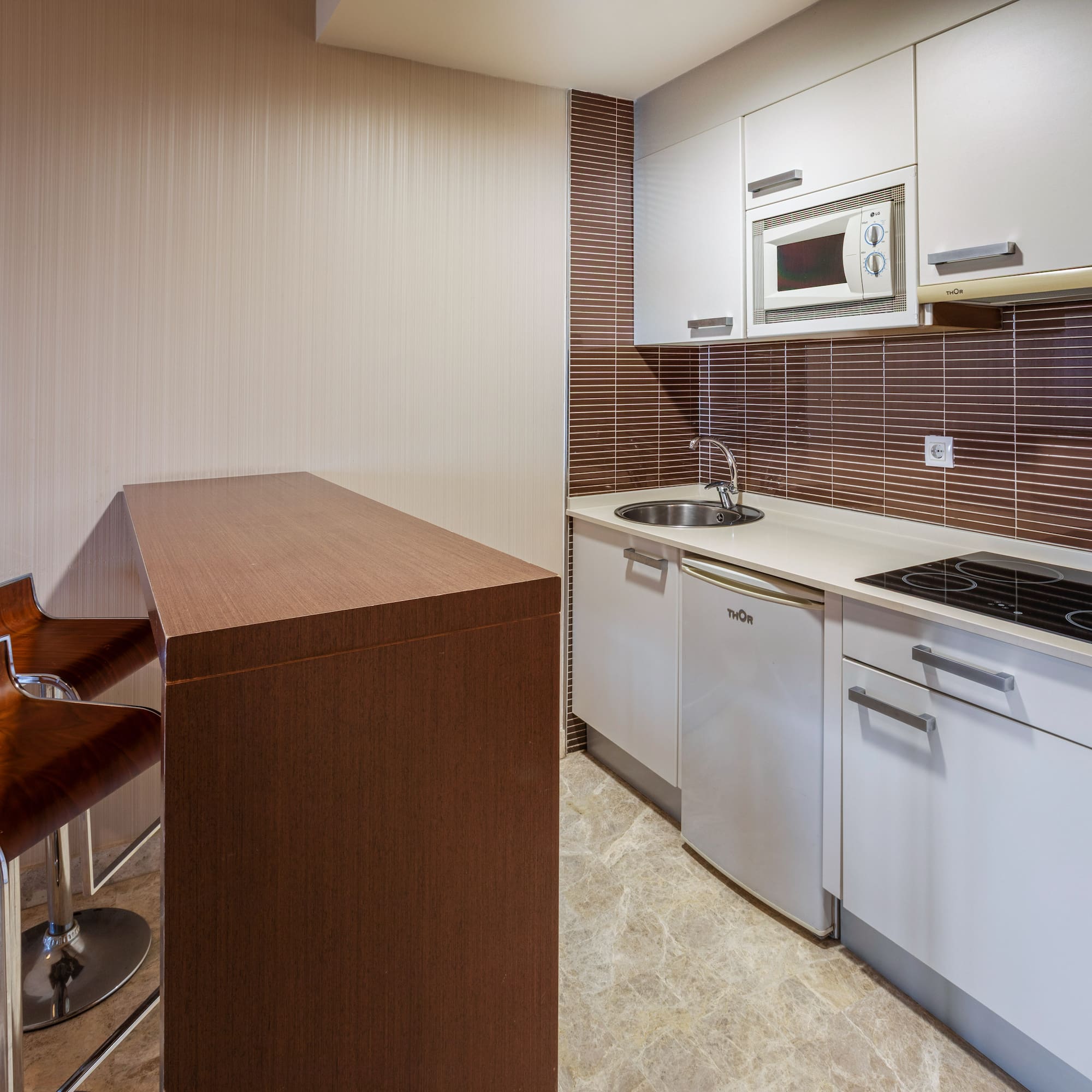 Modern compact kitchen with white cabinets, brown backsplash, and bar stools.