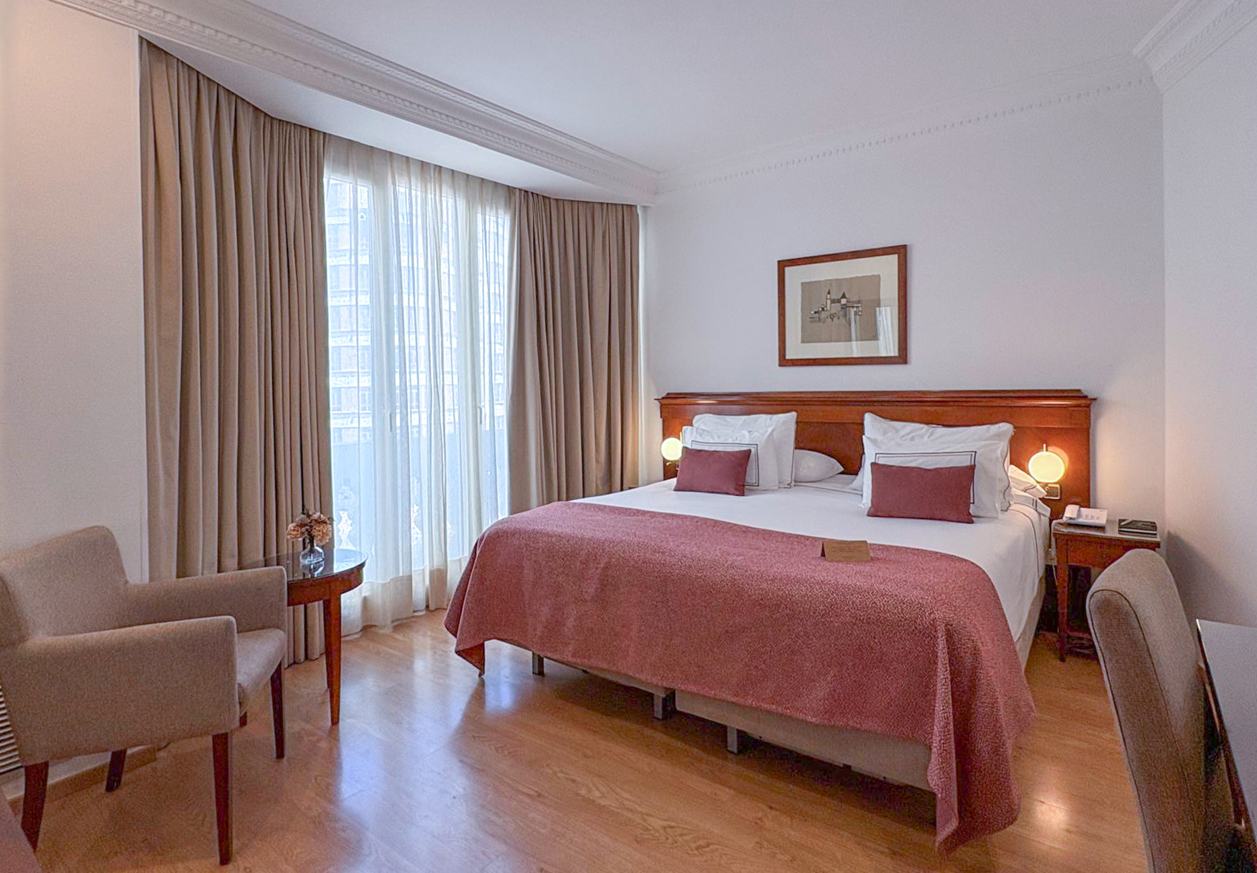 Habitación de hotel con cama grande, cabecero de madera, cortinas beige y parqué.