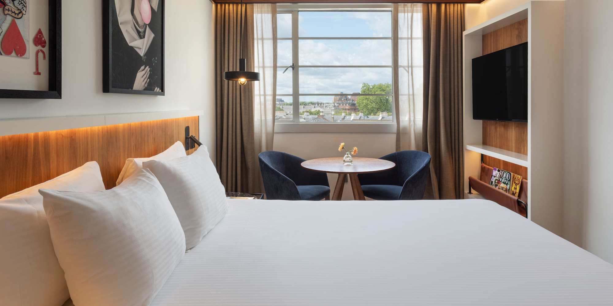 Modern hotel room with white bed, city view window, seating area, and TV.