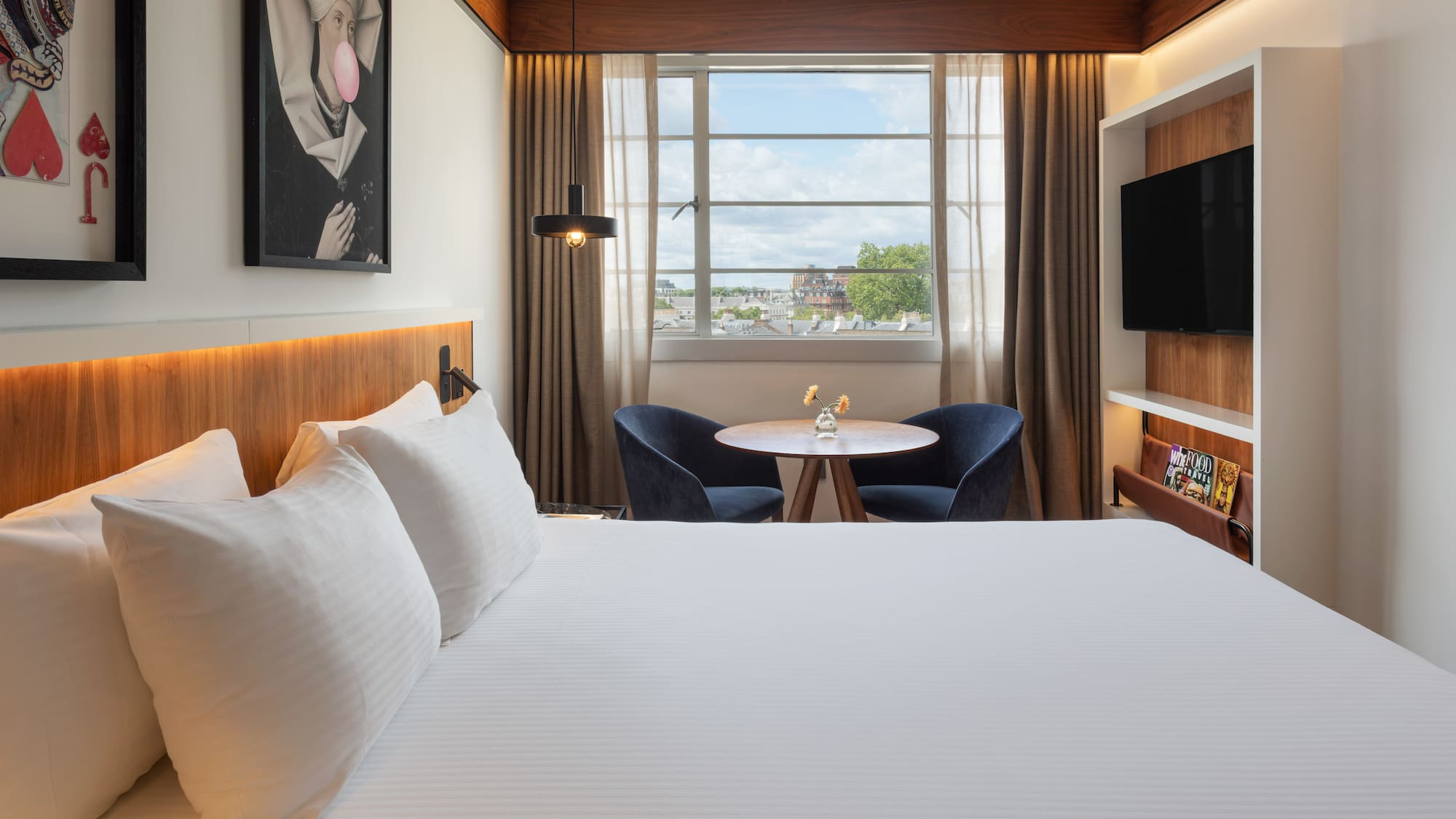 Modern hotel room with white bed, city view window, seating area, and TV.