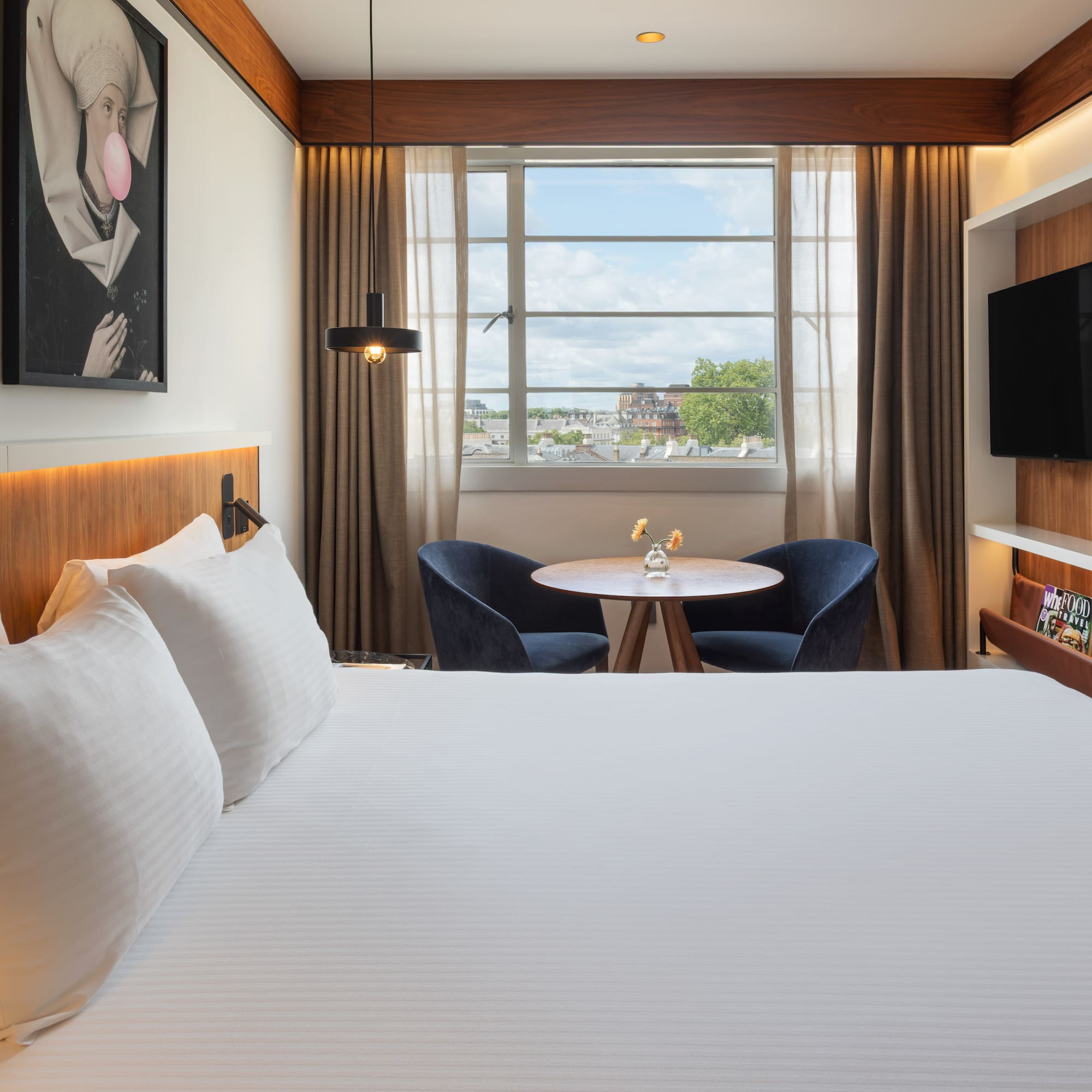 Modern hotel room with white bed, city view window, seating area, and TV.