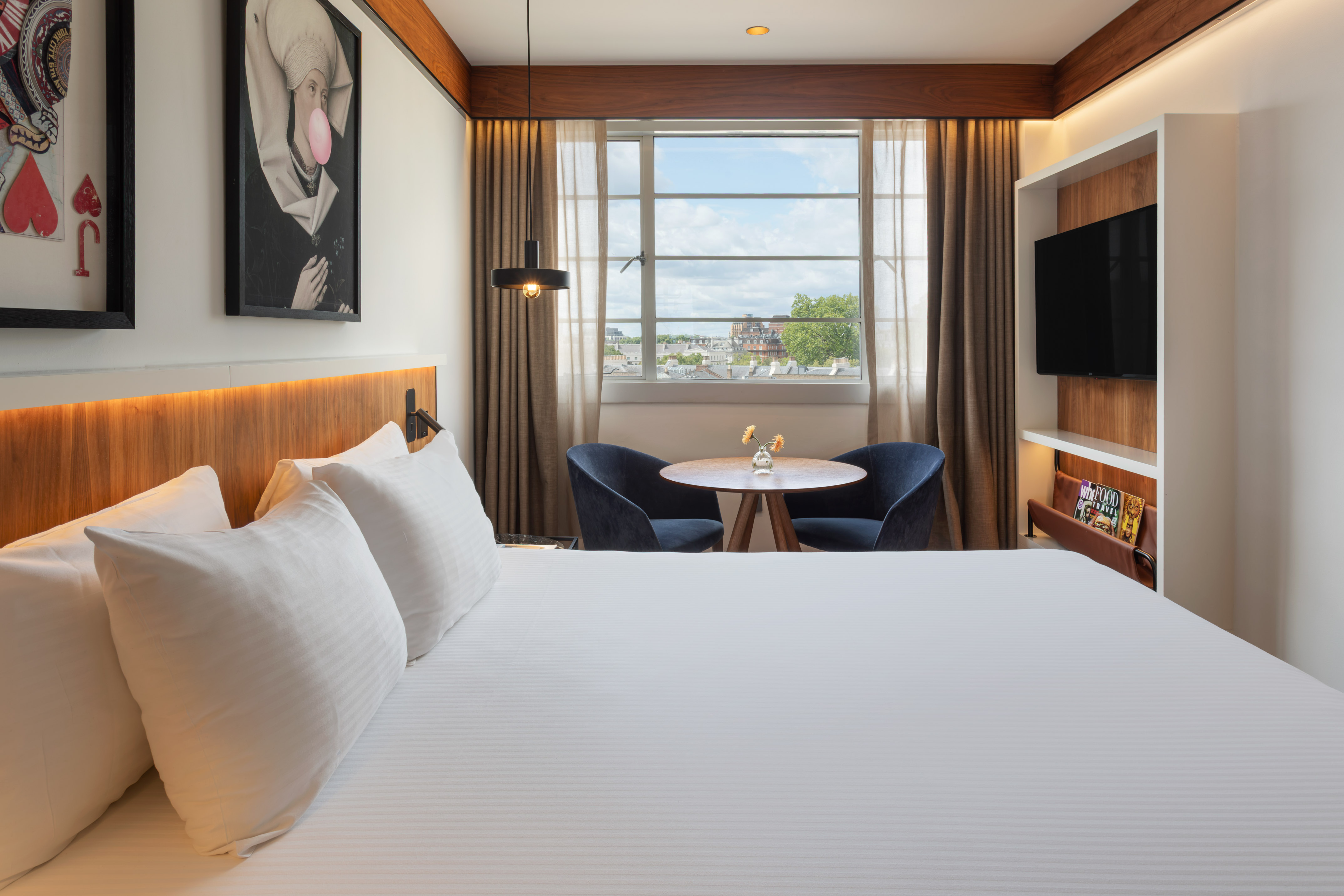 Modern hotel room with white bed, city view window, seating area, and TV.