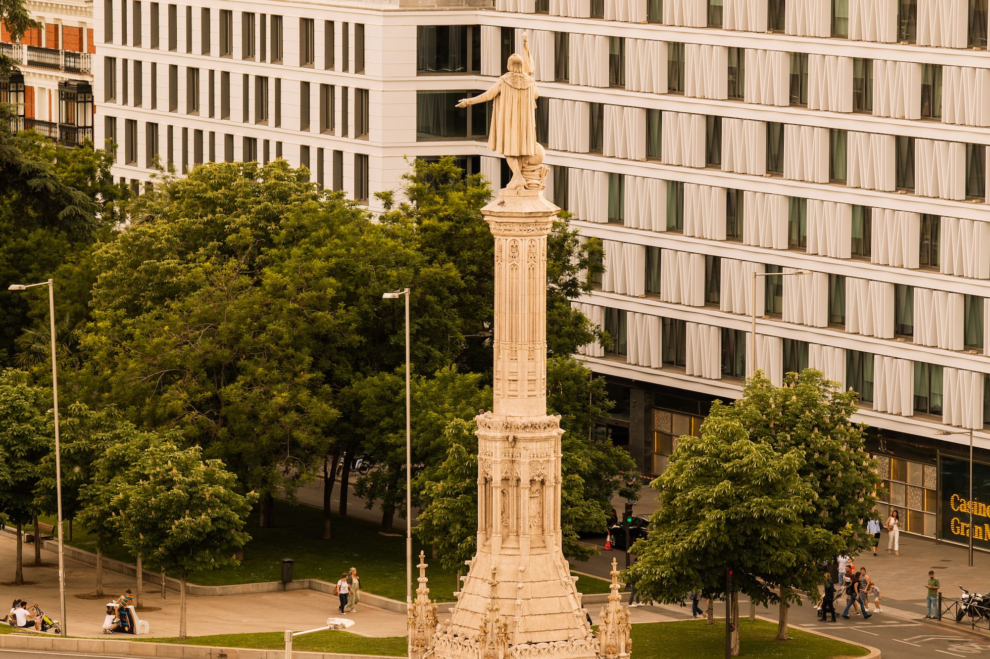 Columbus Monument in Madrid, with fountain, green trees, and city buildings.