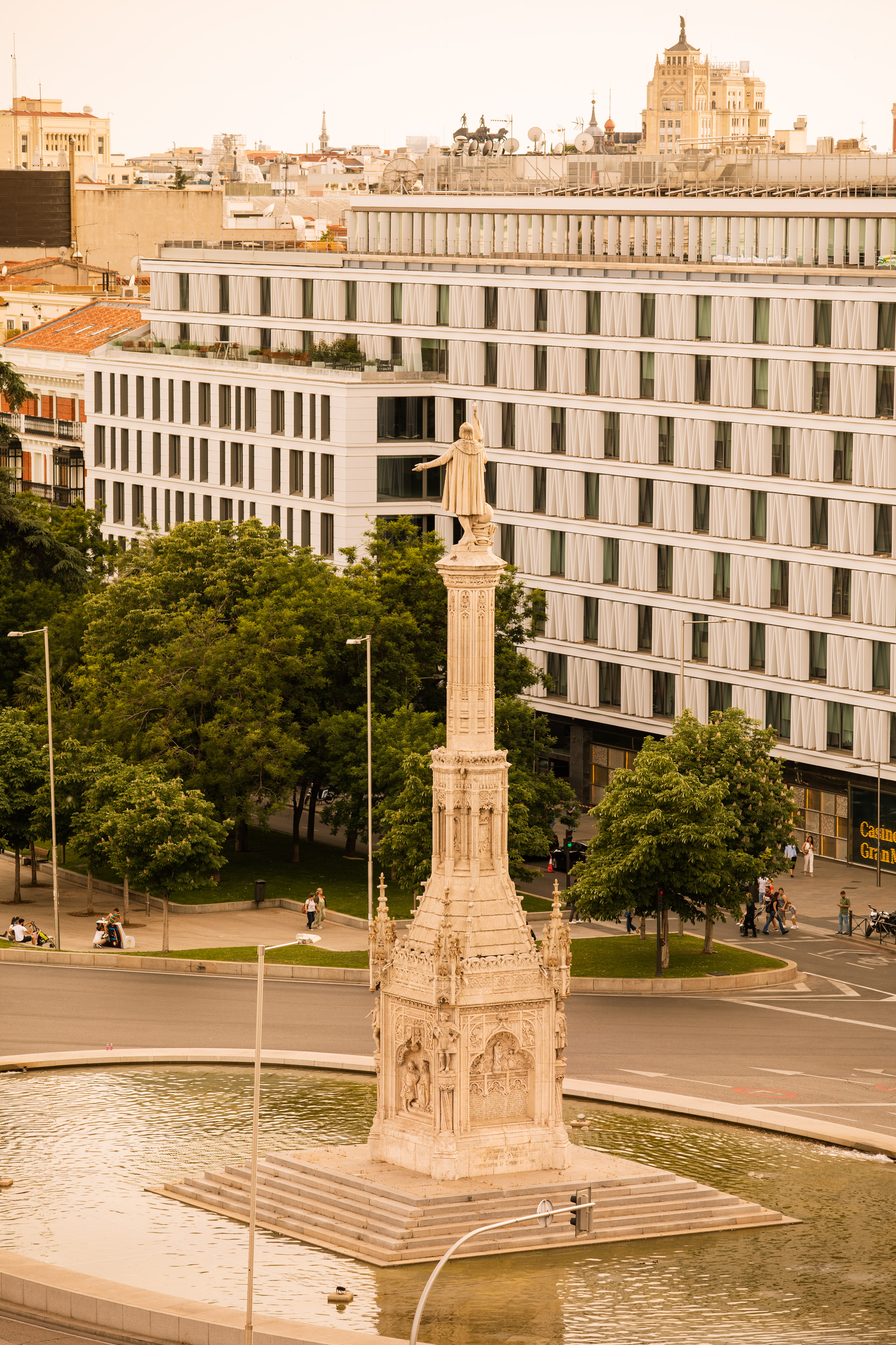 Columbus Monument in Madrid, with fountain, green trees, and city buildings.