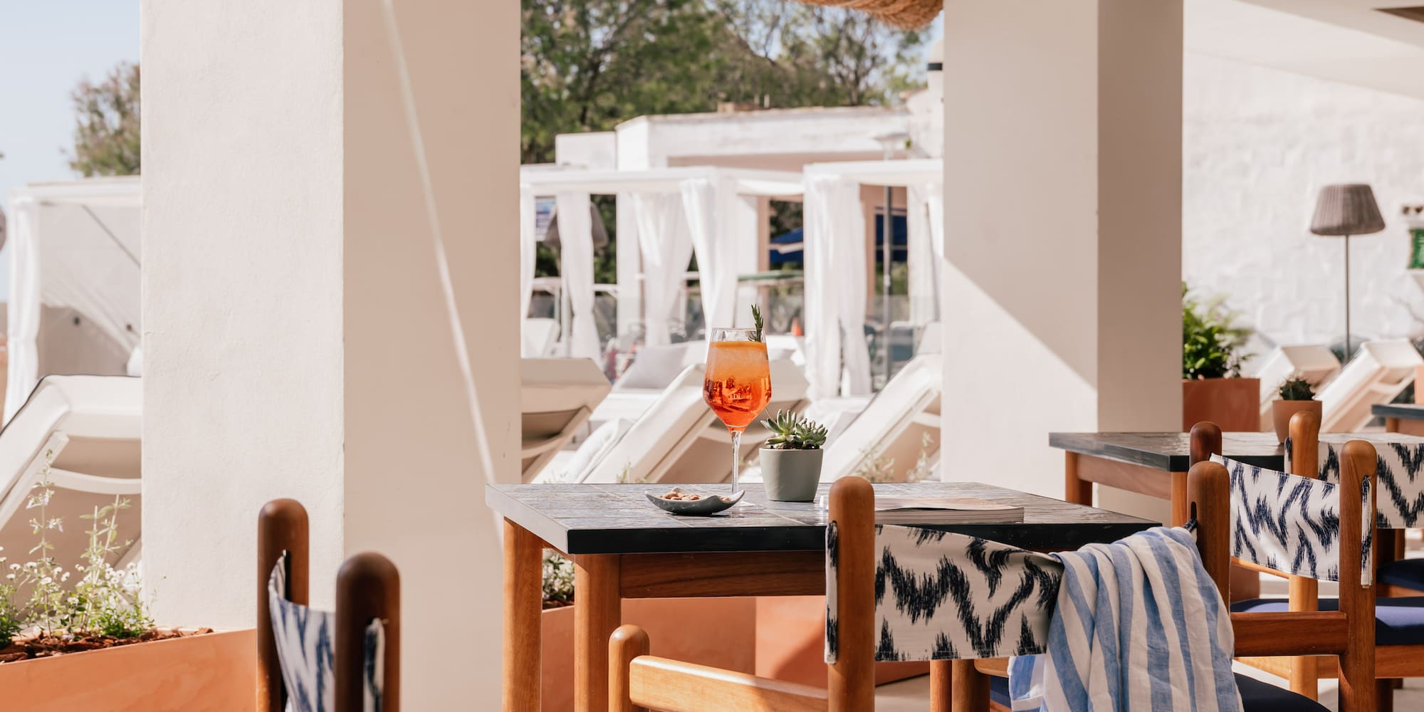 Sunny hotel patio with orange cocktail on wooden table and blue chairs.