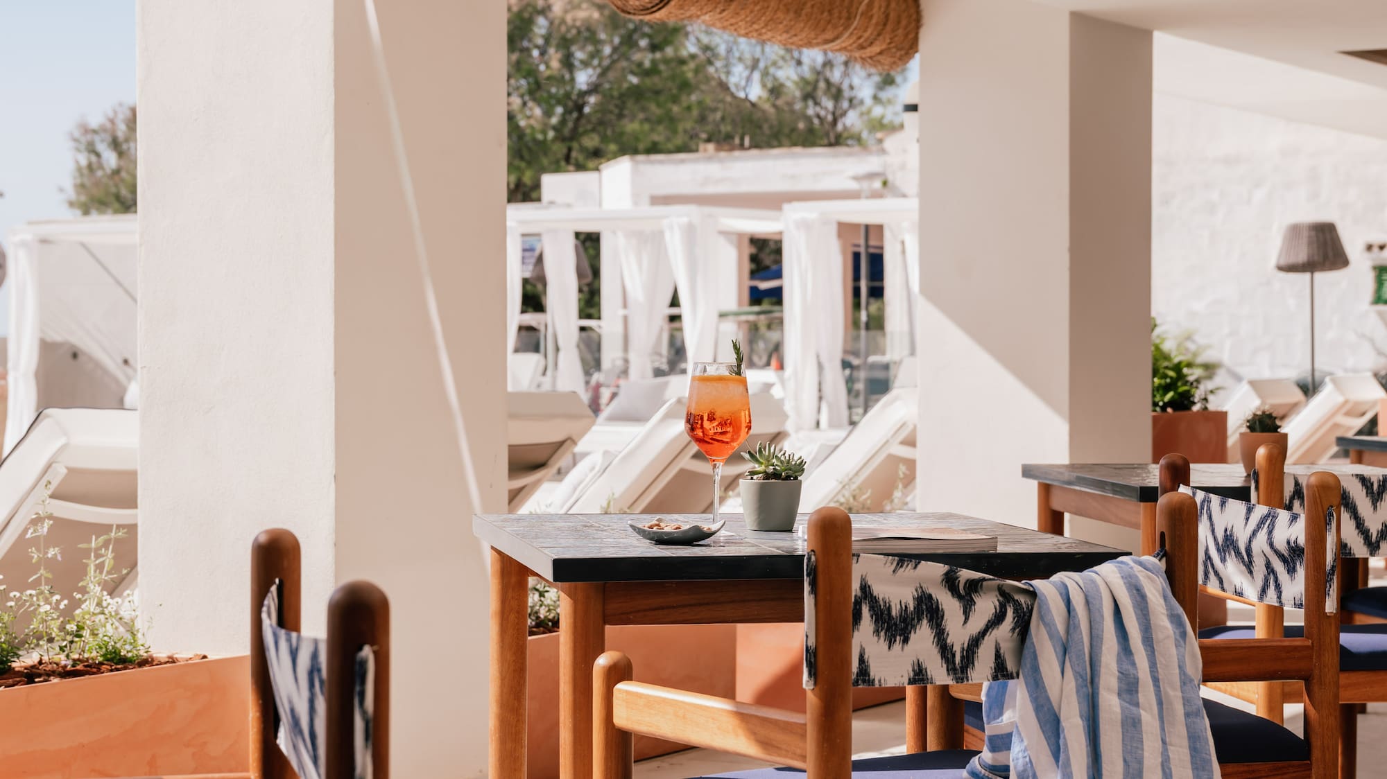 Sunny hotel patio with orange cocktail on wooden table and blue chairs.