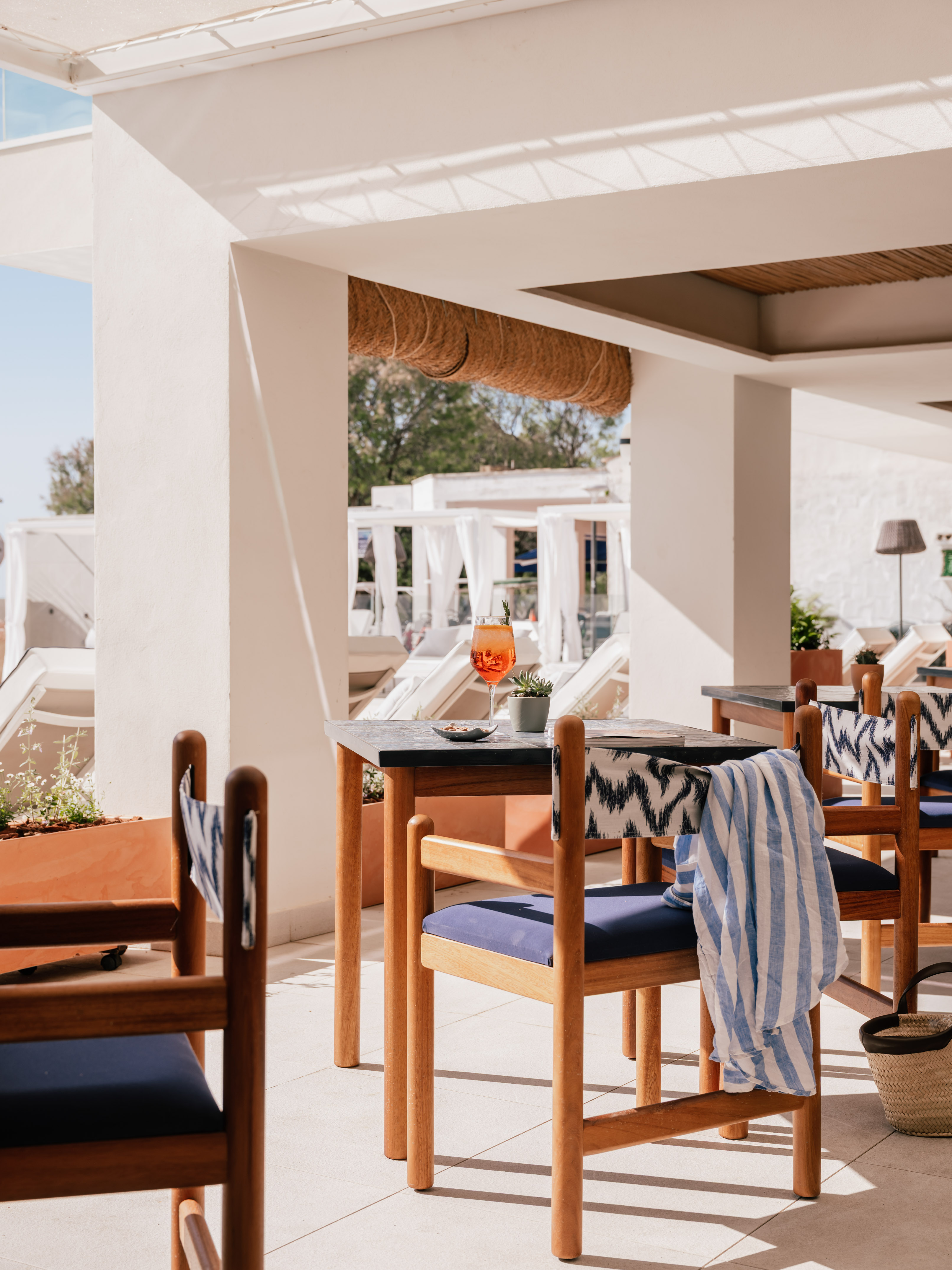 Sunny hotel patio with orange cocktail on wooden table and blue chairs.