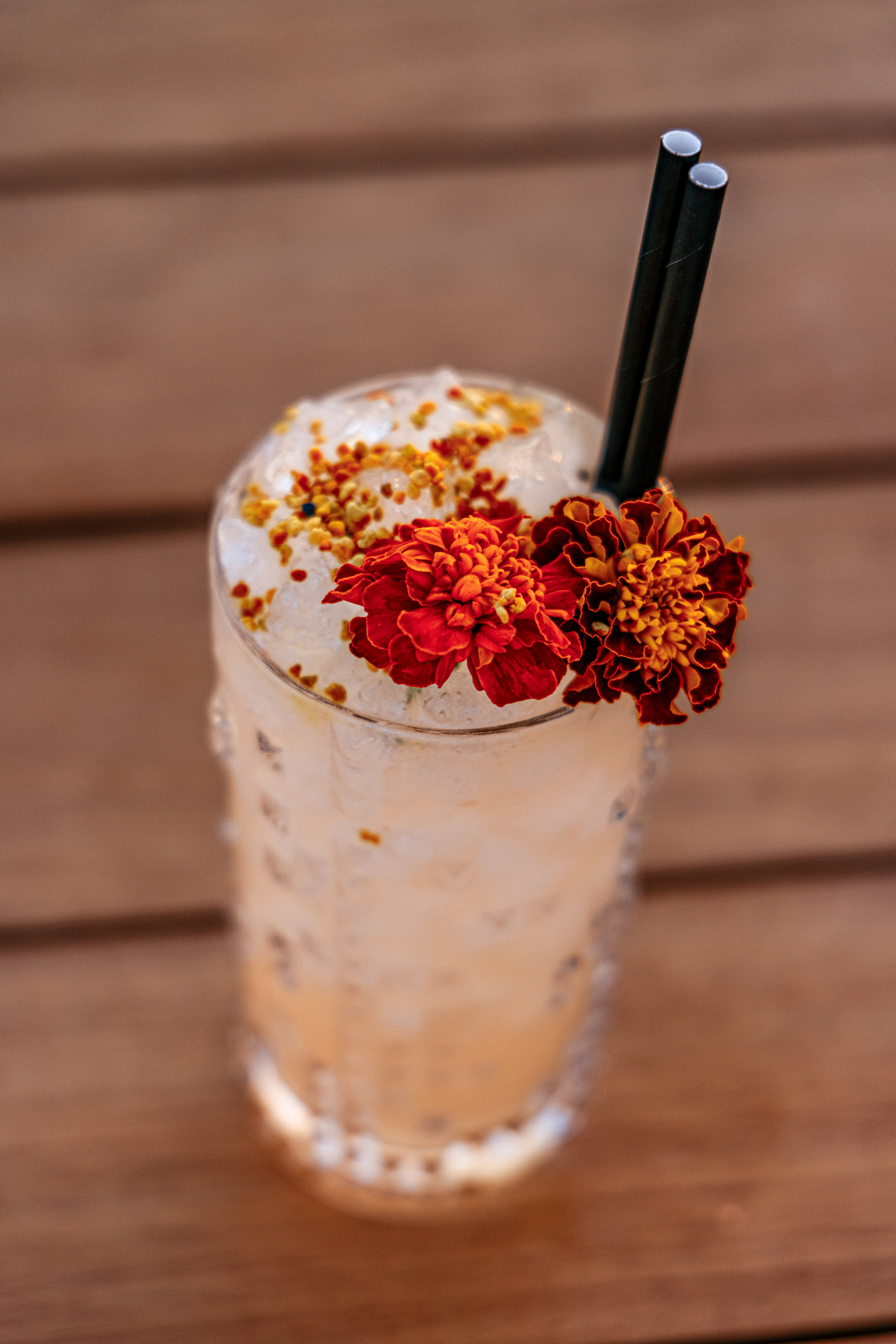 Iced cocktail with marigold flowers, bee pollen, and black straws on wood.