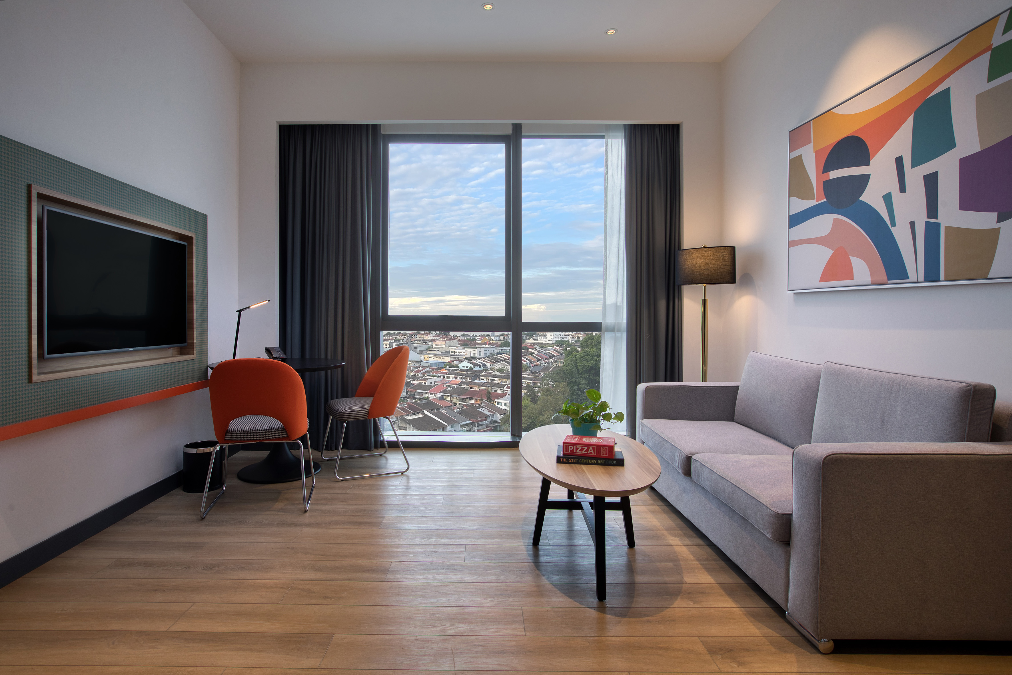 Sleek hotel room interior with city view, grey sofa, TV, and orange accent chairs.