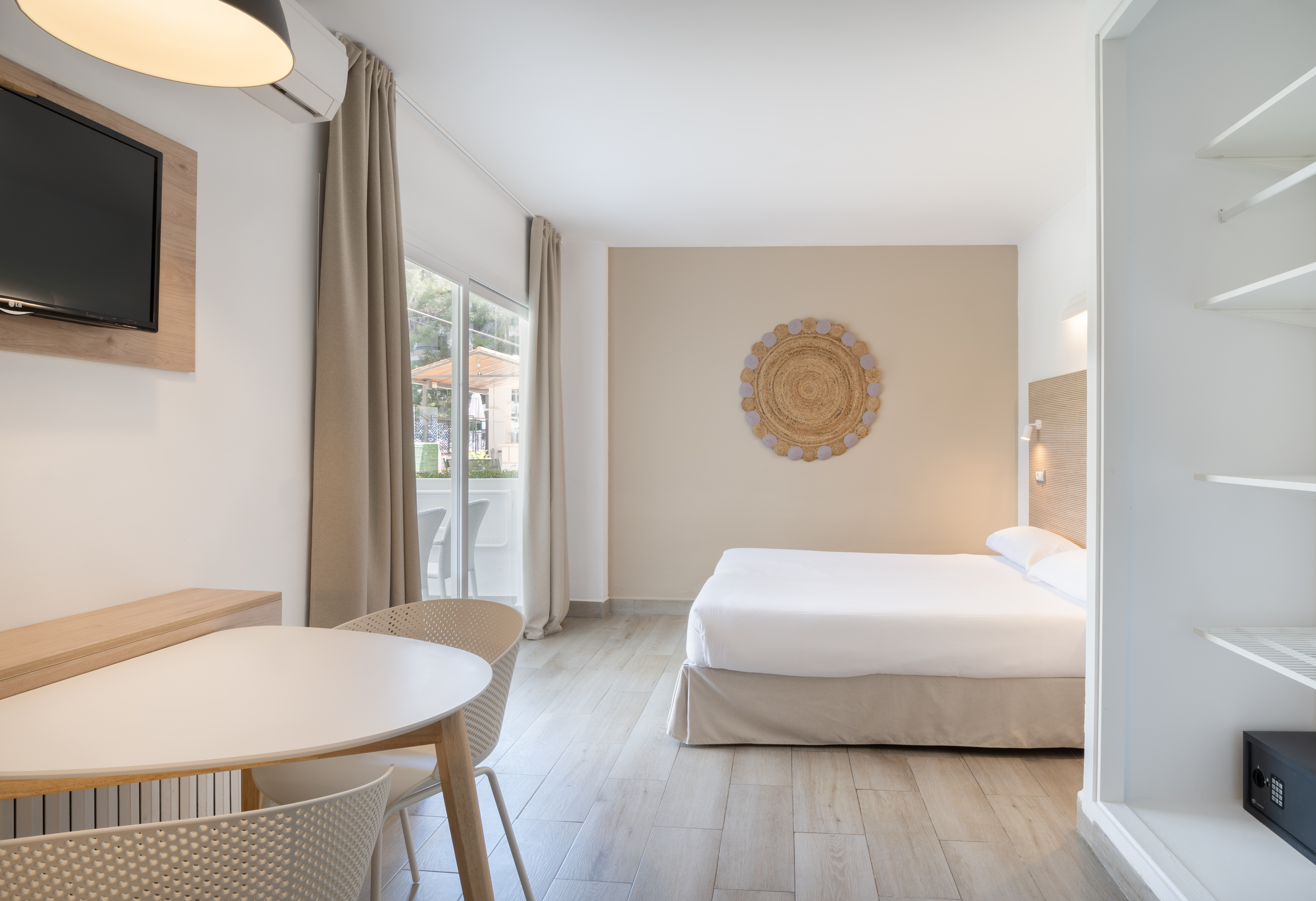 Modern hotel studio with bed, dining table, TV, and bright balcony access.