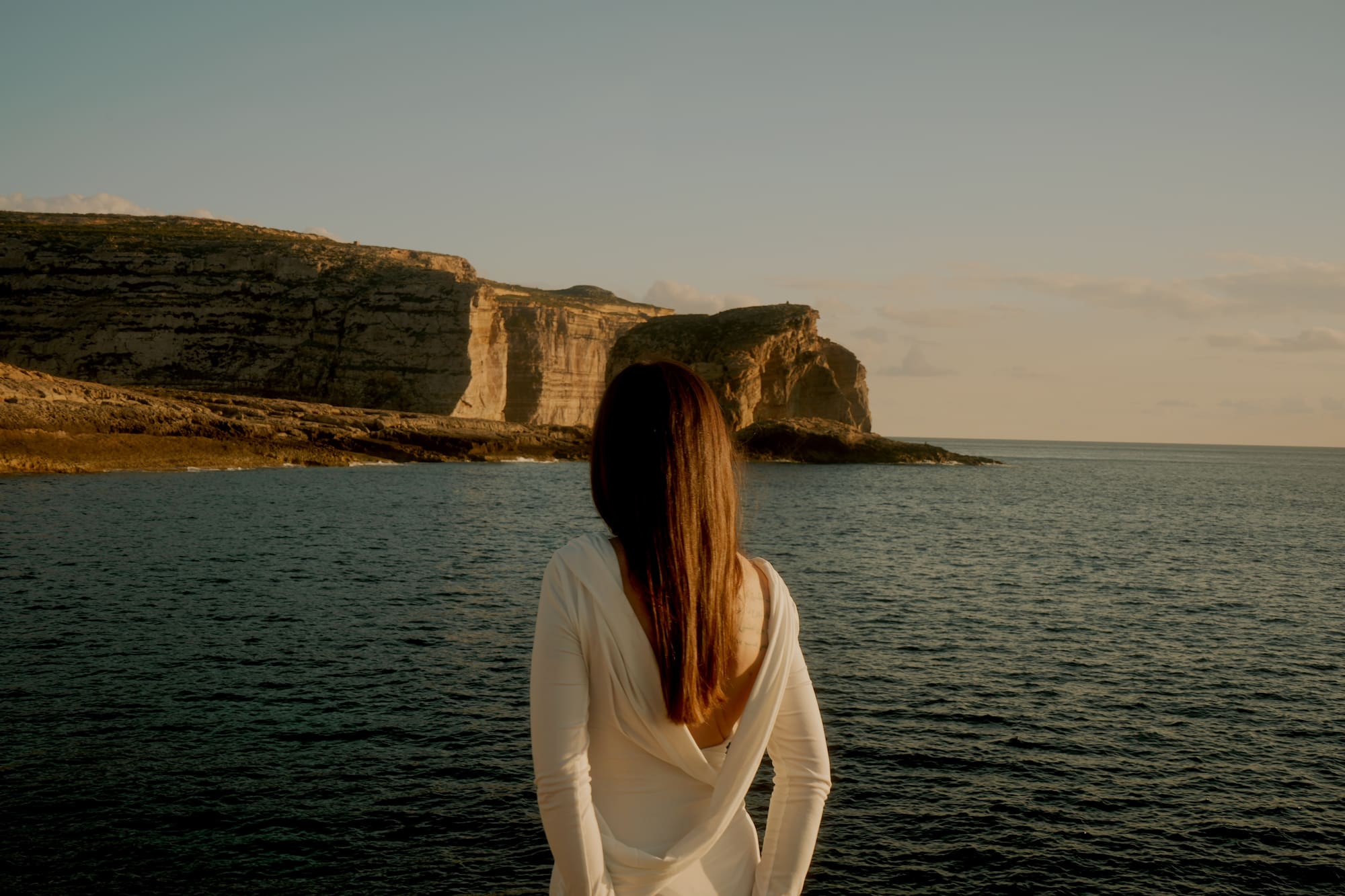 Woman in white dress looking at dramatic sea cliffs at sunset.