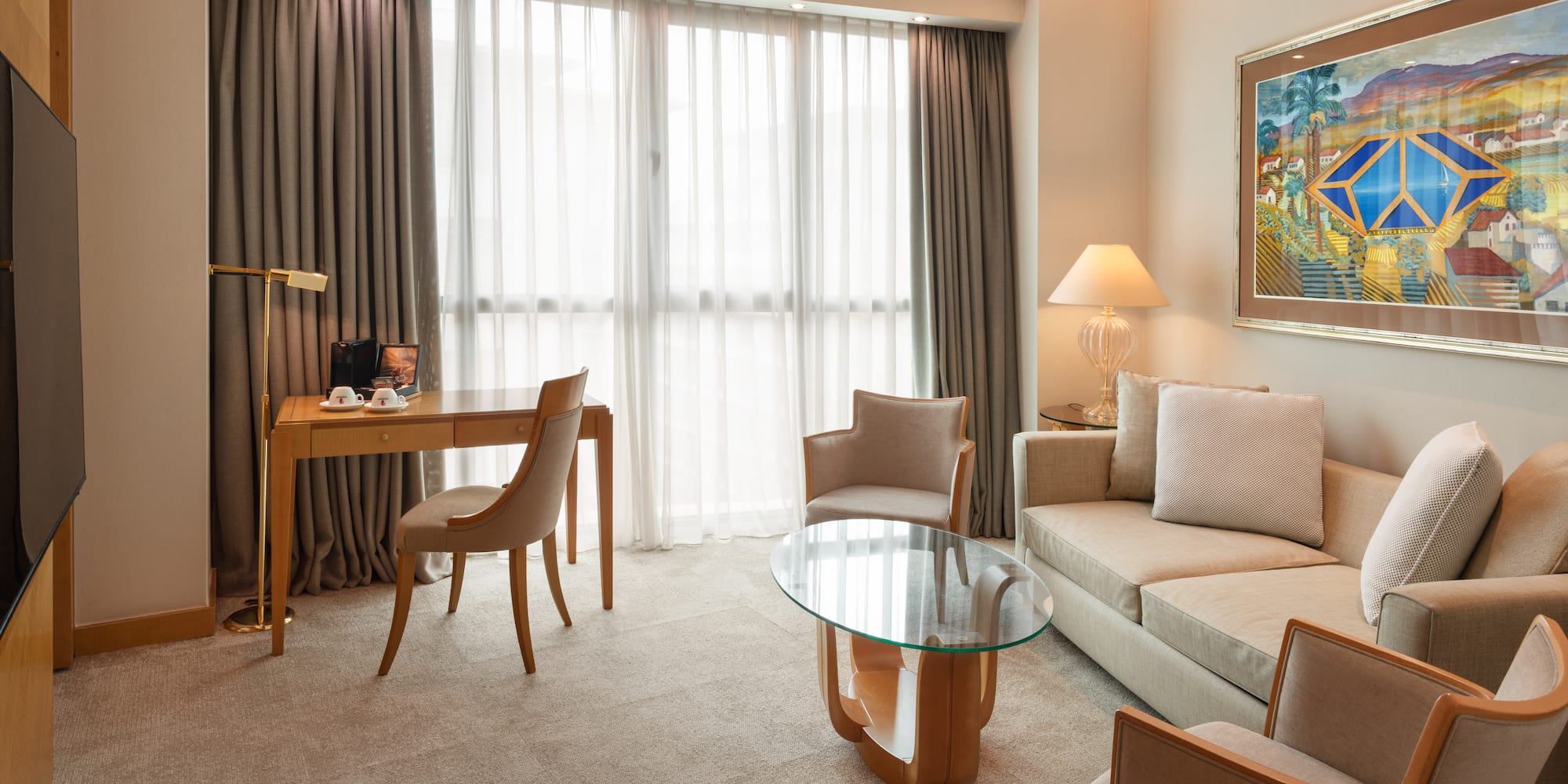 Modern hotel suite living area with sofa, desk, armchairs, and large window.