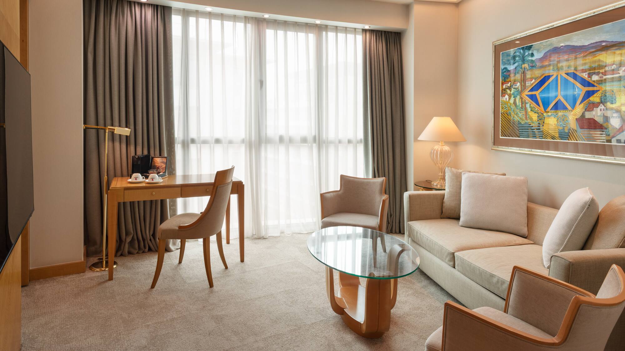 Modern hotel suite living area with sofa, desk, armchairs, and large window.