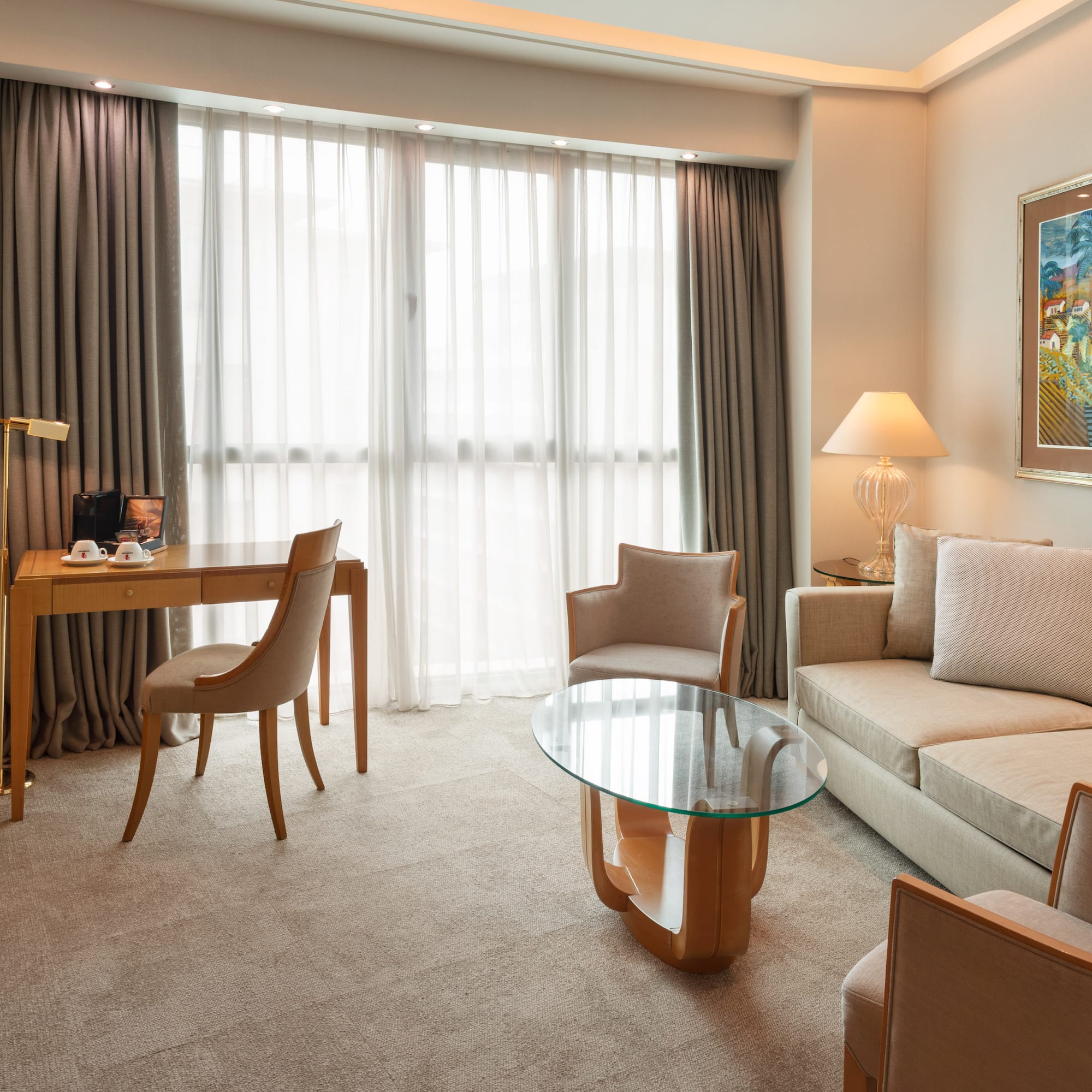 Modern hotel suite living area with sofa, desk, armchairs, and large window.