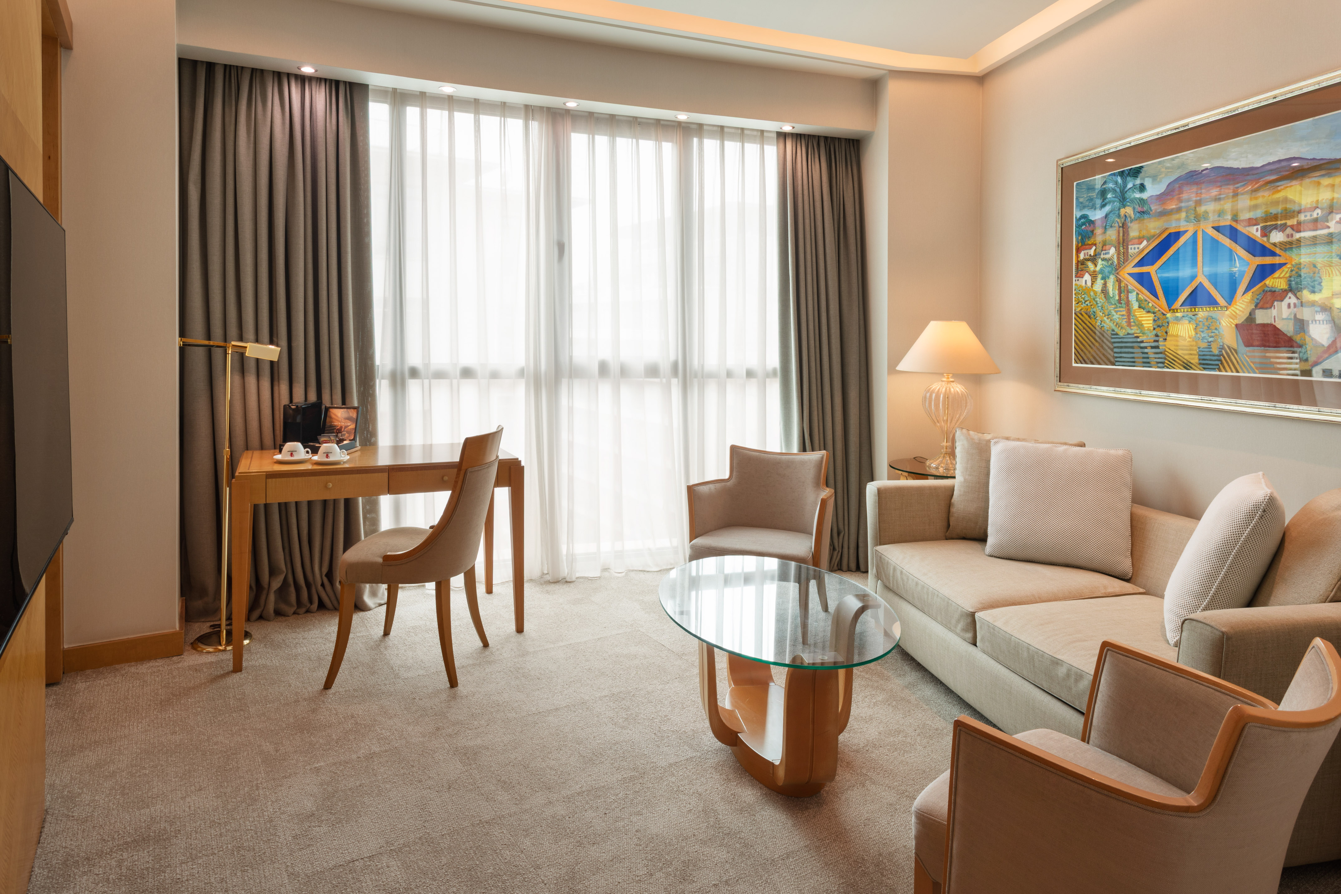 Modern hotel suite living area with sofa, desk, armchairs, and large window.