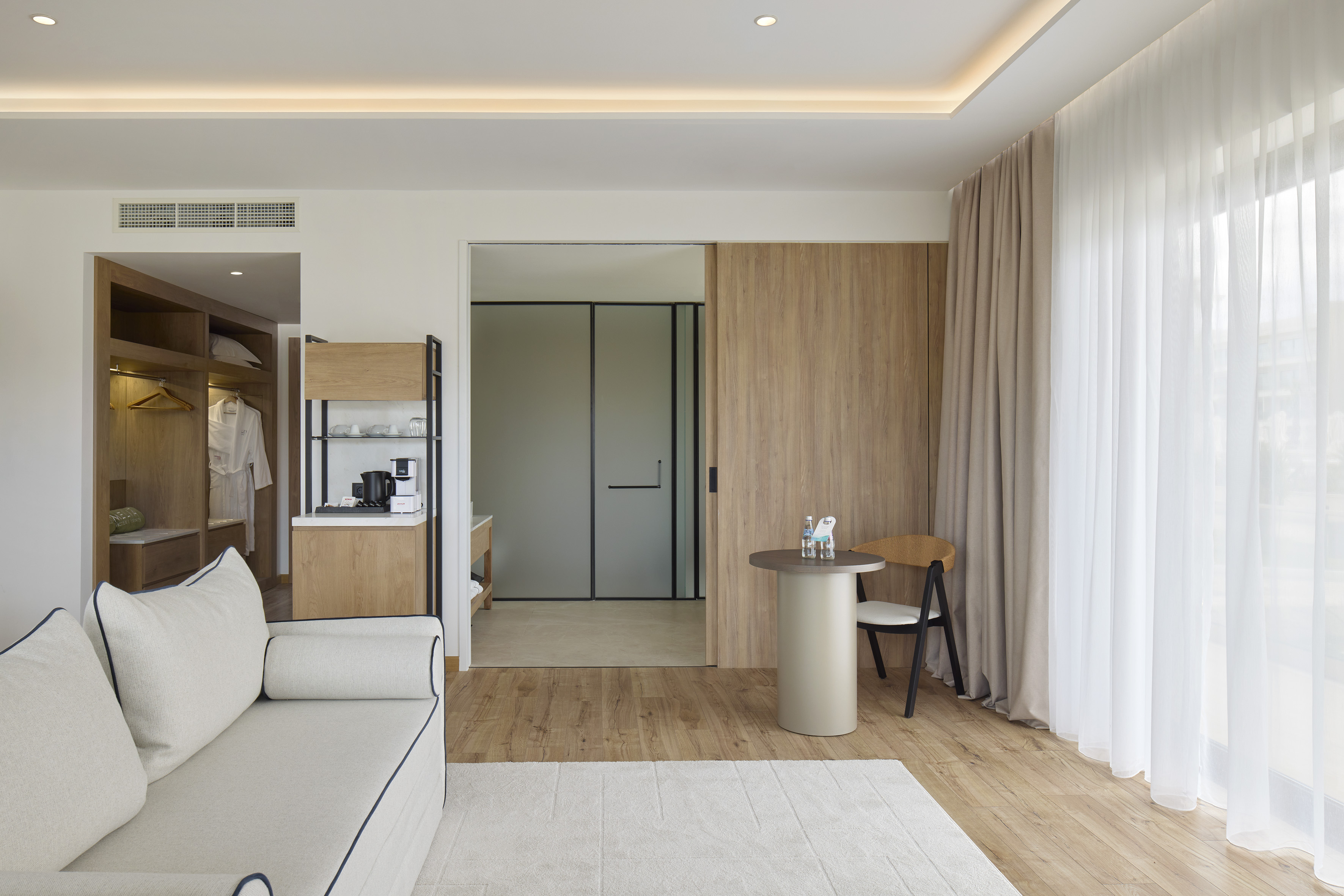 Modern hotel suite with sofa, coffee bar, wood floors, and large window.
