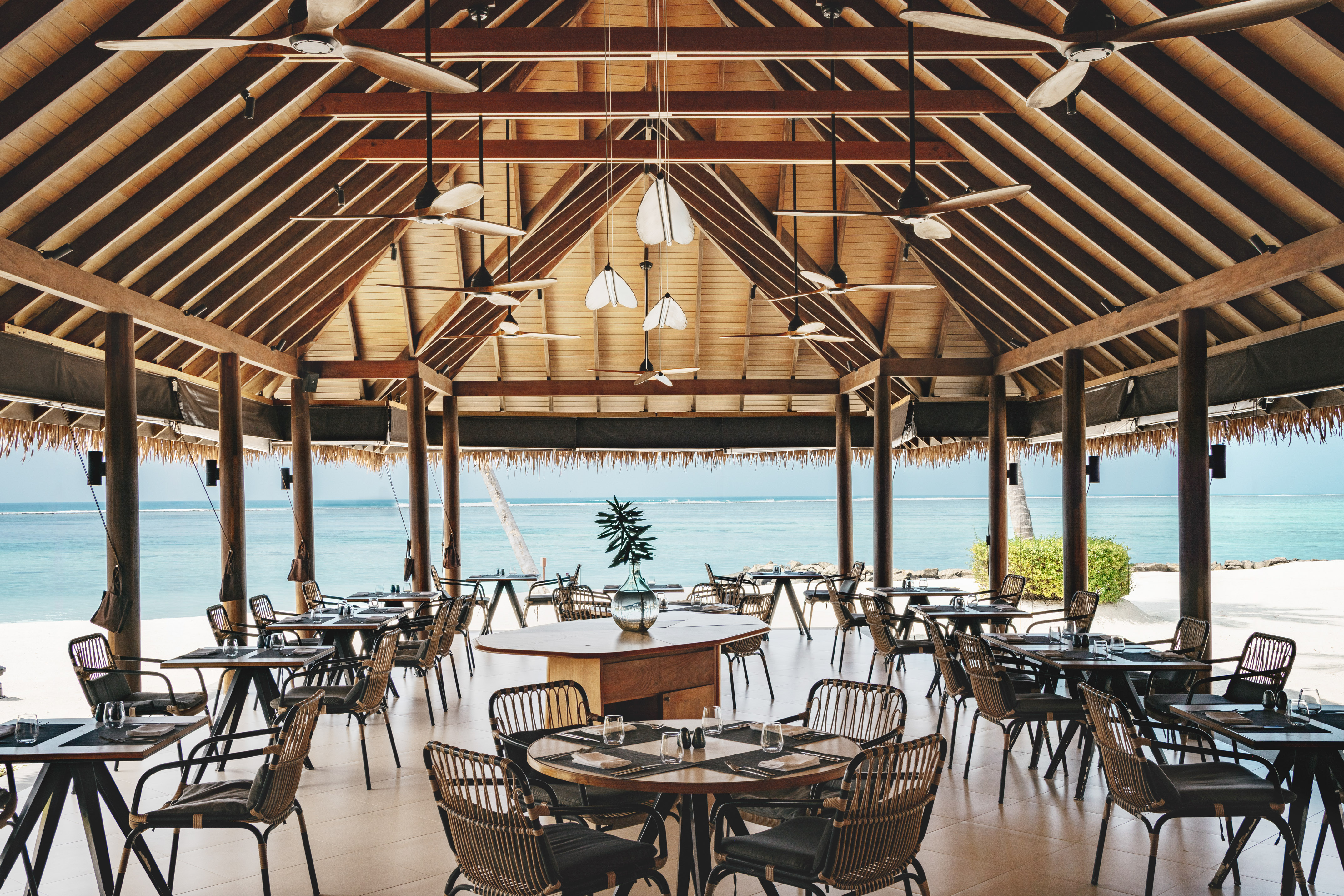 Tropical open-air beach restaurant with wooden roof, fans, ocean view.