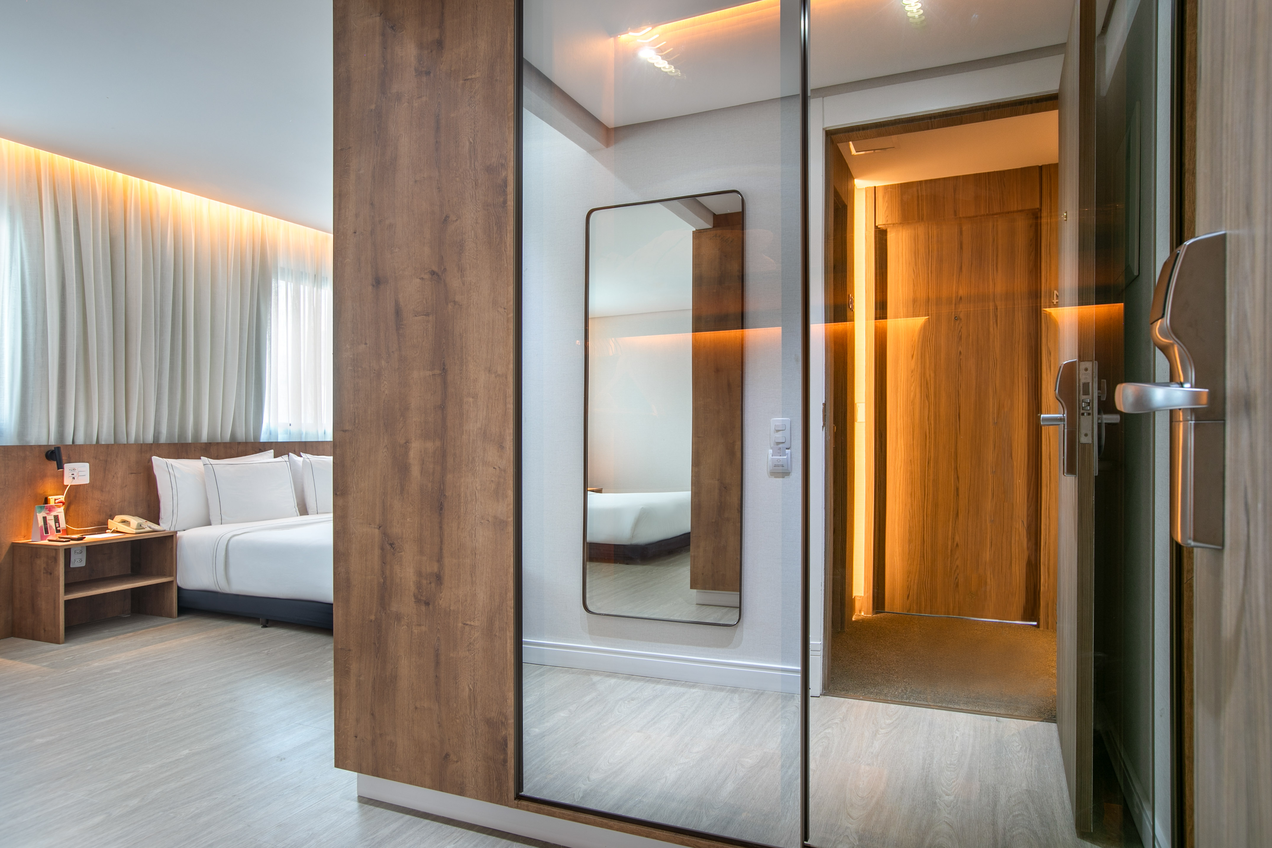 Modern hotel room with bed, light wood, large mirror, and warm strip lighting.