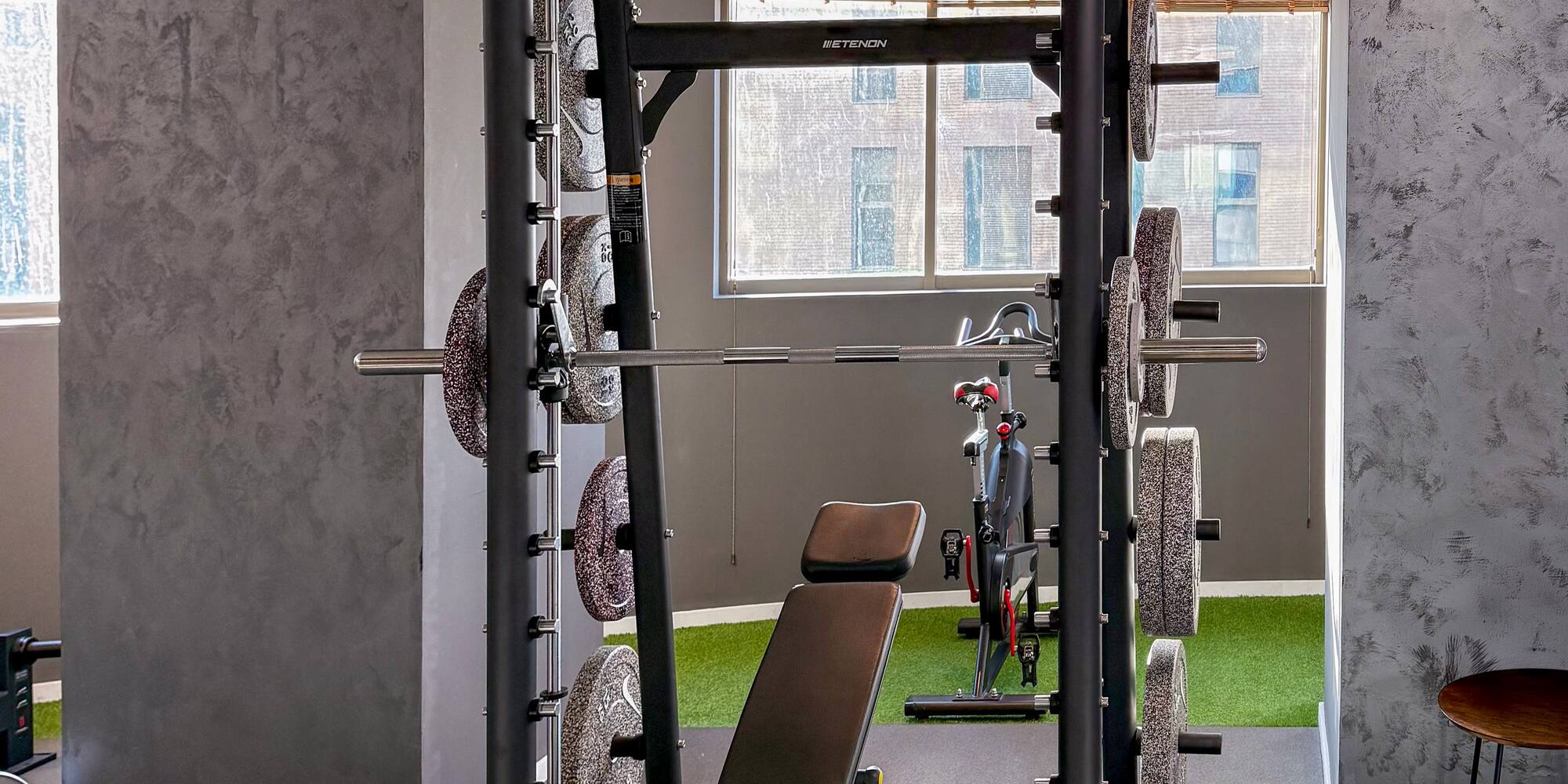 Modern gym with Smith machine, weights, bench, and urban window view.