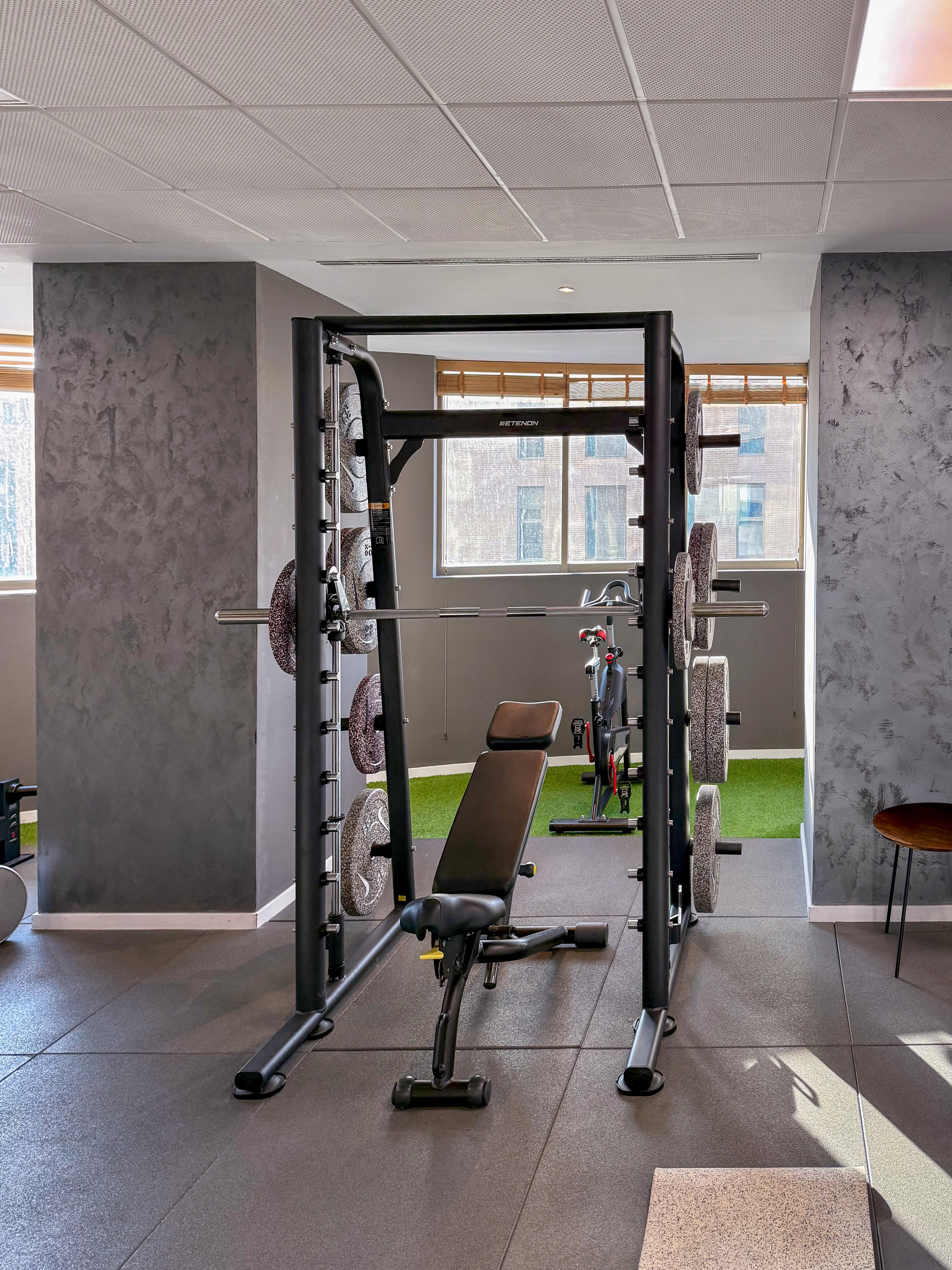 Modern gym with Smith machine, weights, bench, and urban window view.