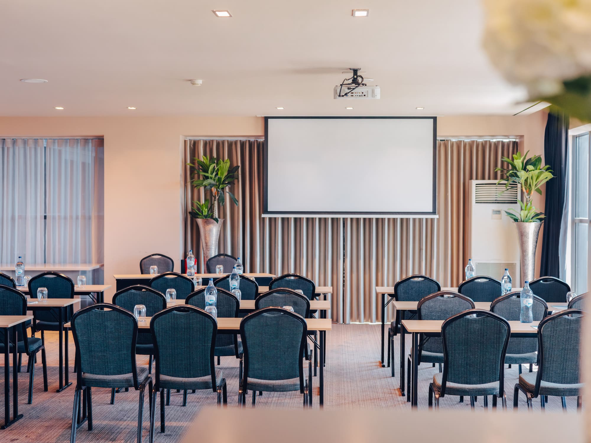 Conference room setup with projector screen, tables, chairs, and bottled water.
