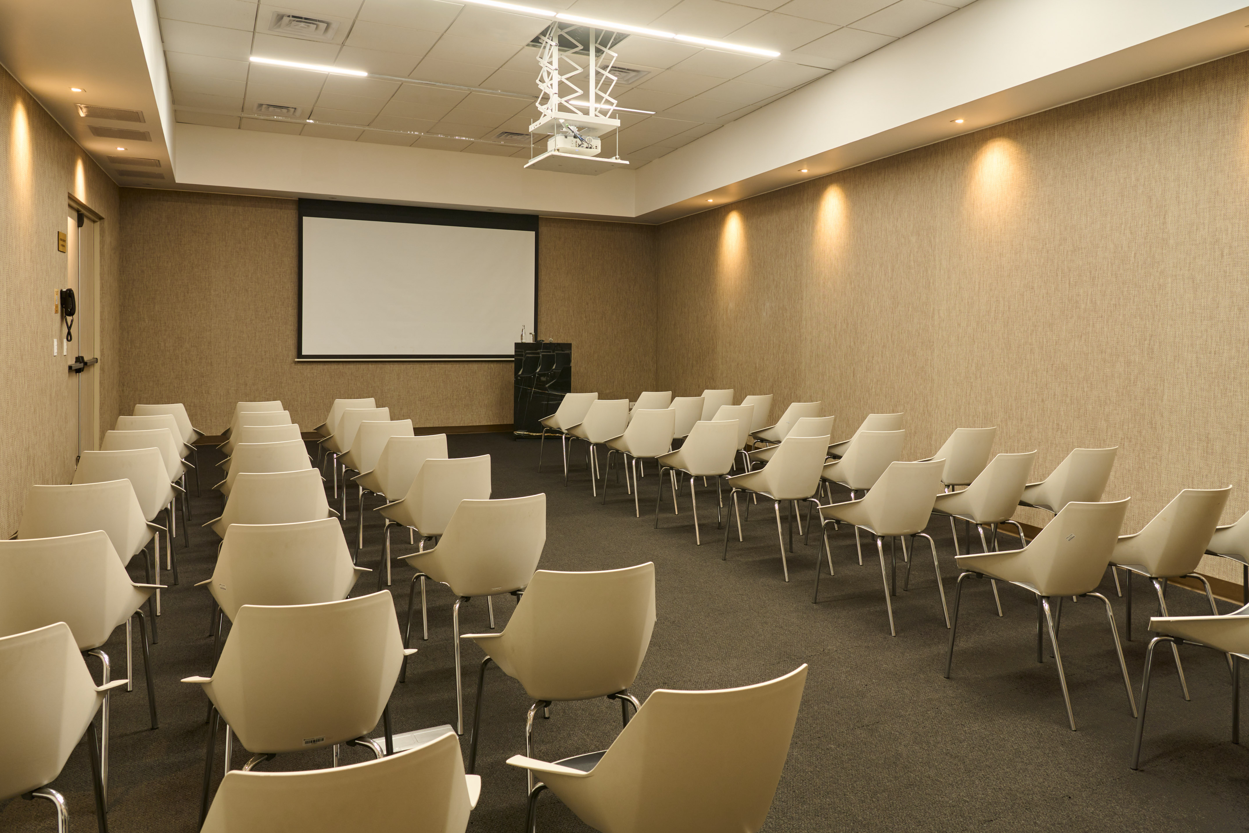 Empty conference room with rows of modern chairs, projector screen, and podium.
