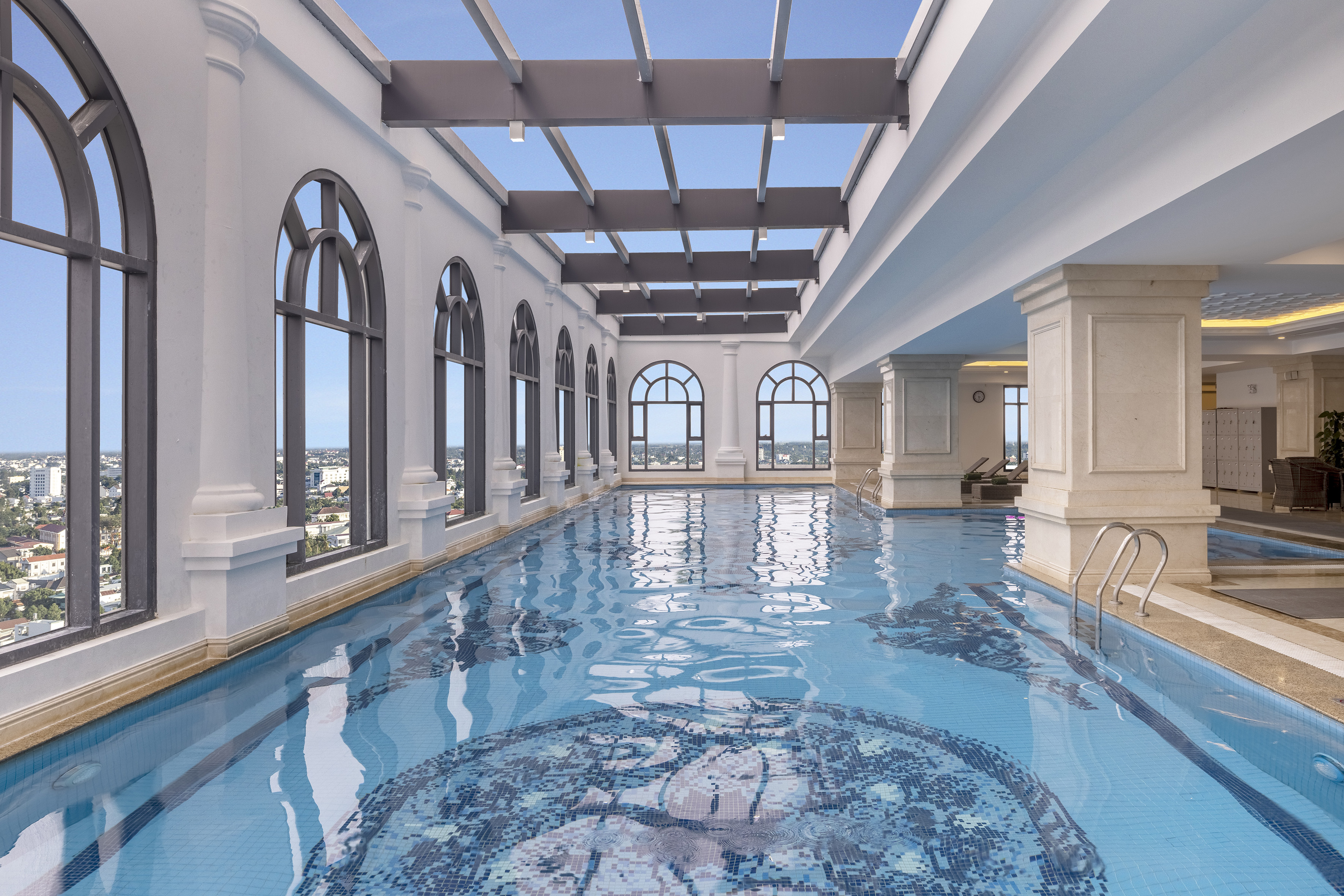 Indoor swimming pool with city views through arched windows and skylight ceiling.
