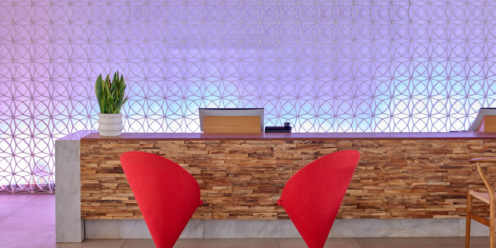 Hotel reception with wood counter, two red chairs, and geometric backlit wall.