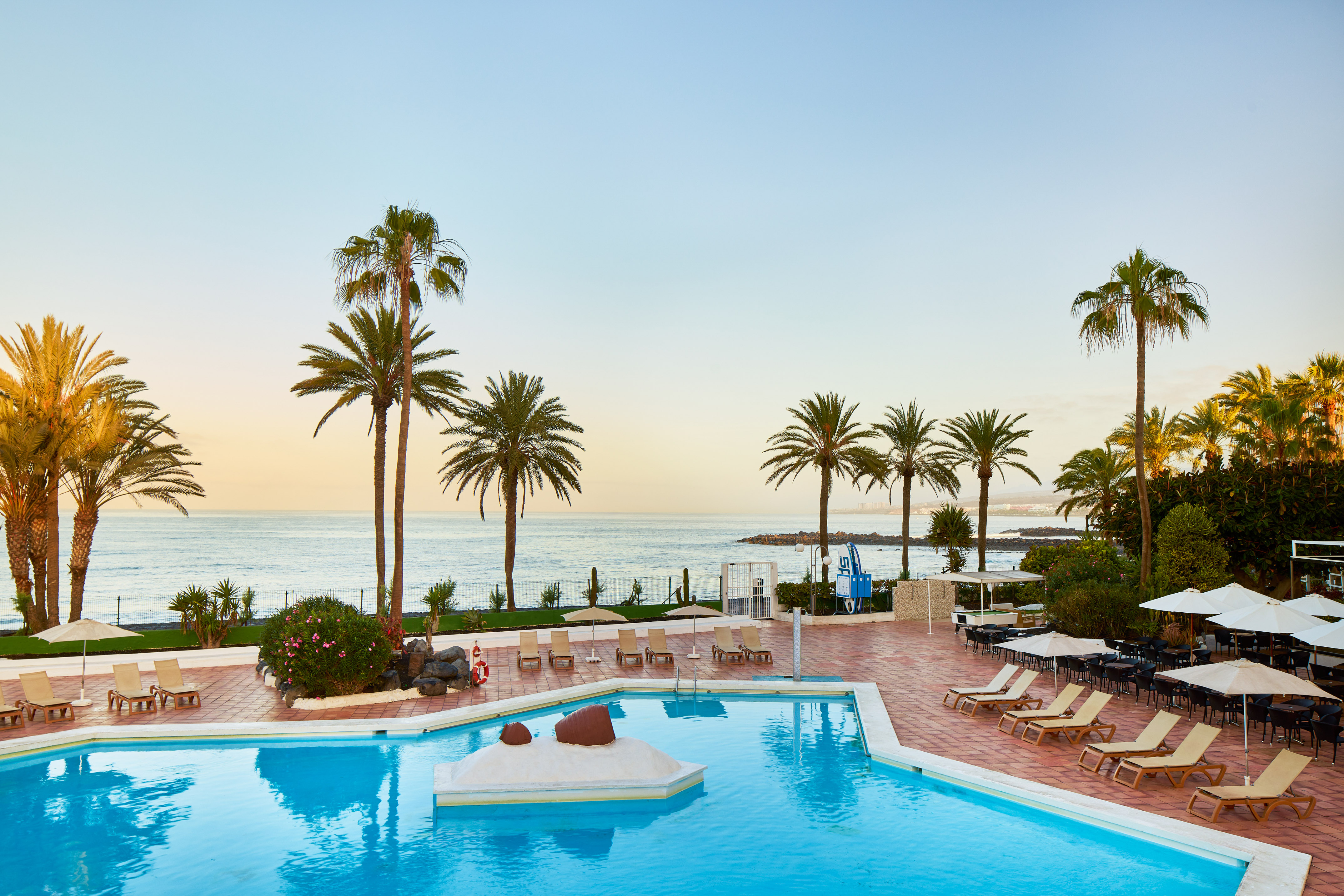 Tropical resort pool with palm trees, ocean view, and sun loungers at sunset.