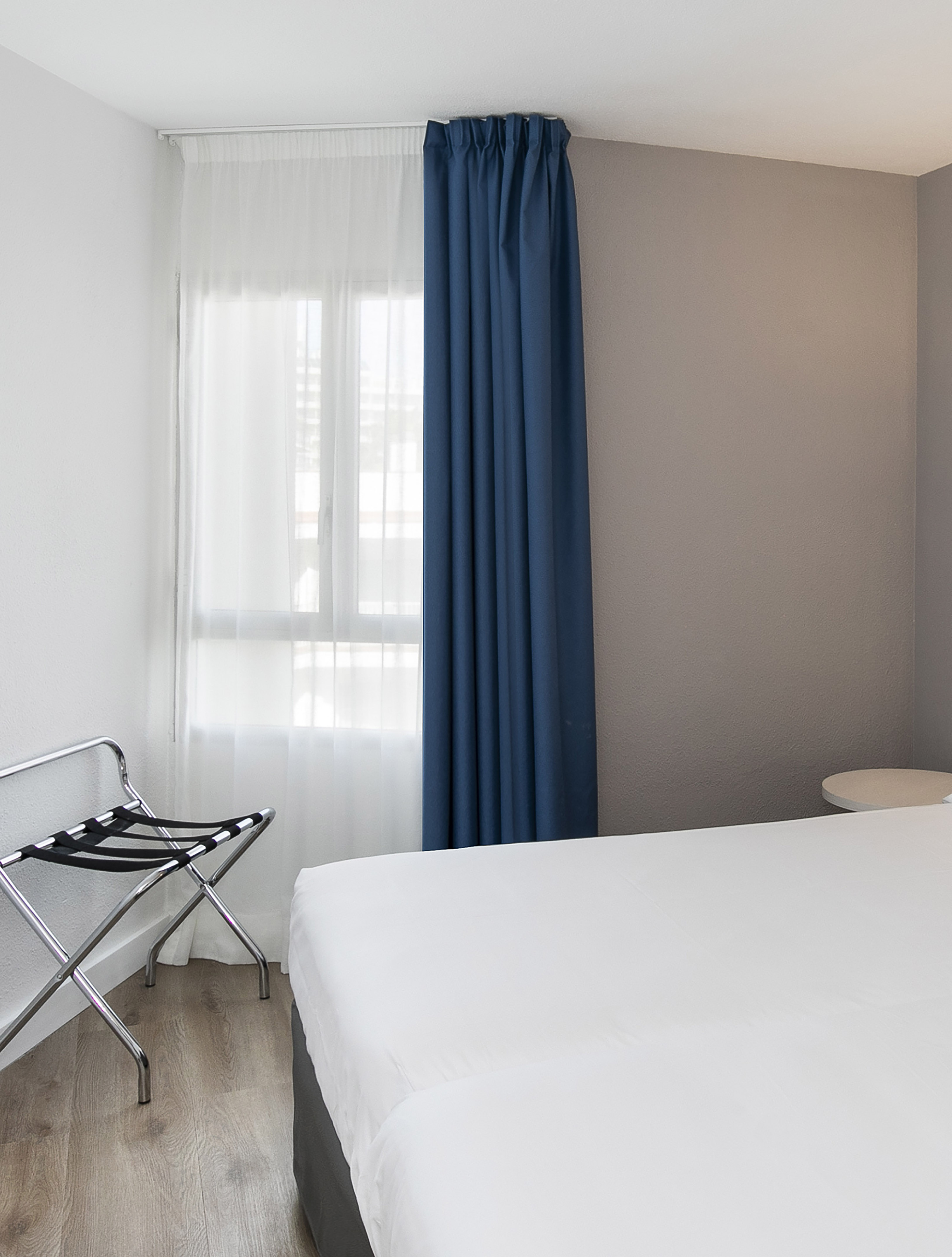 Bright bedroom with white bed, blue curtains, and chrome luggage rack by window.