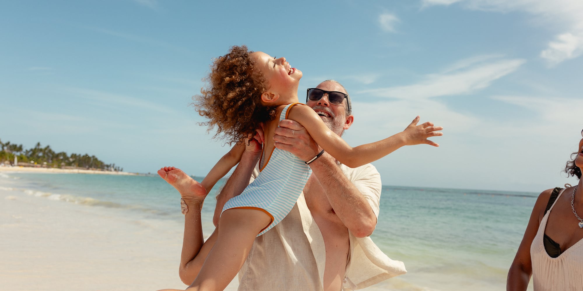 Smiling man lifts happy child high on a sunny beach by the ocean.