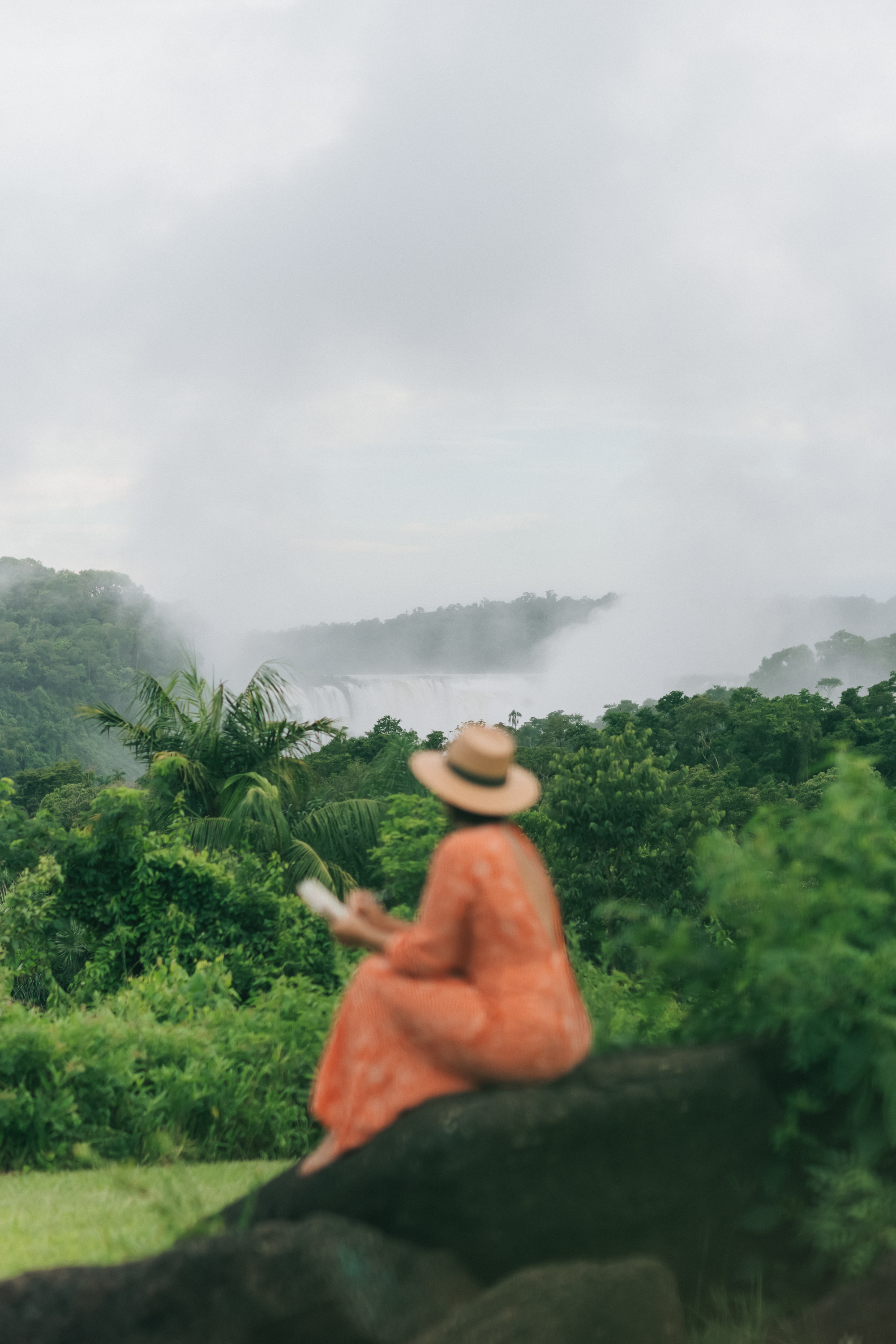 Woman in hat observing misty tropical jungle waterfall.