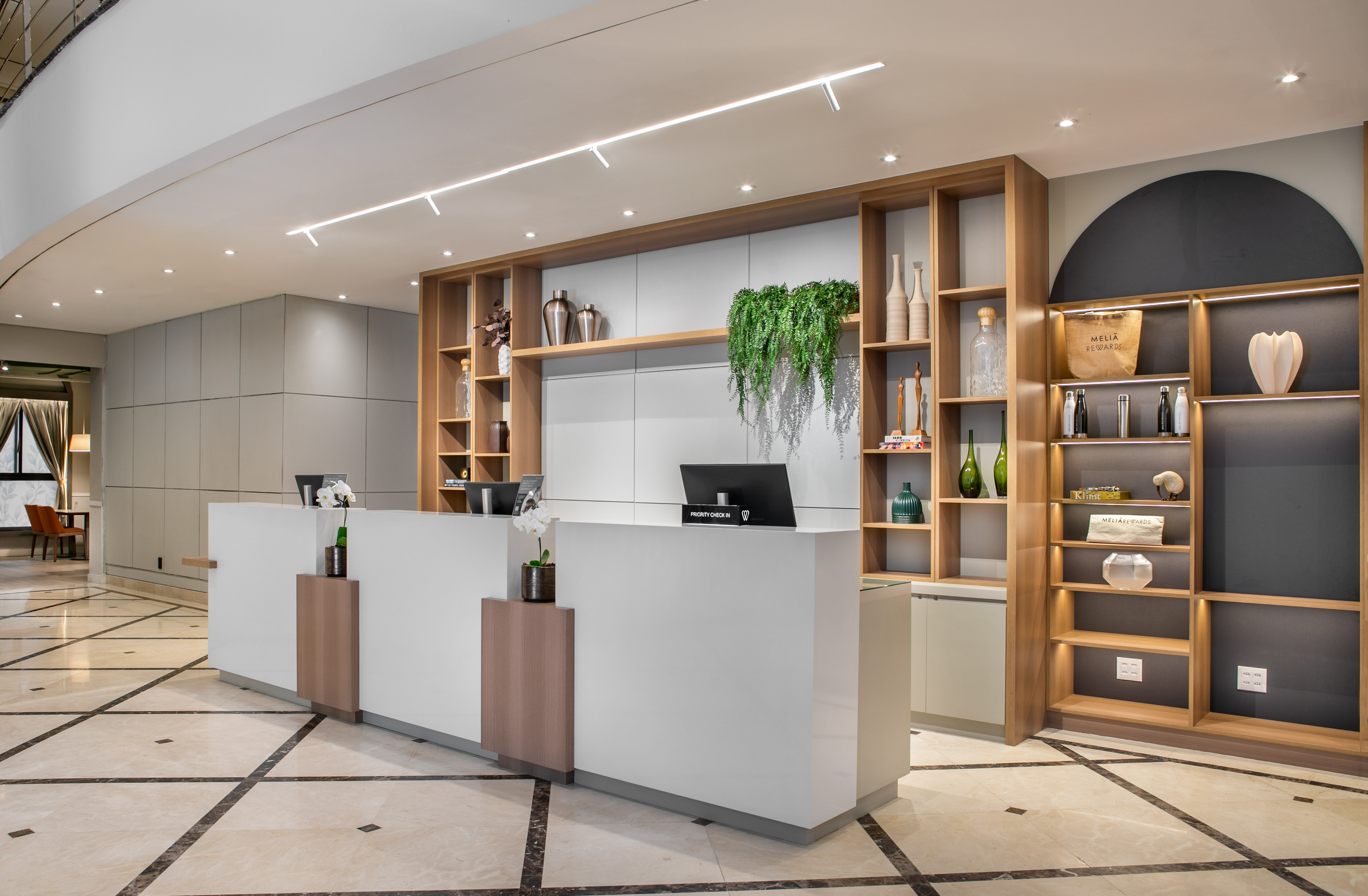 Modern hotel reception desk with wood shelves, green plants, and marble floor.