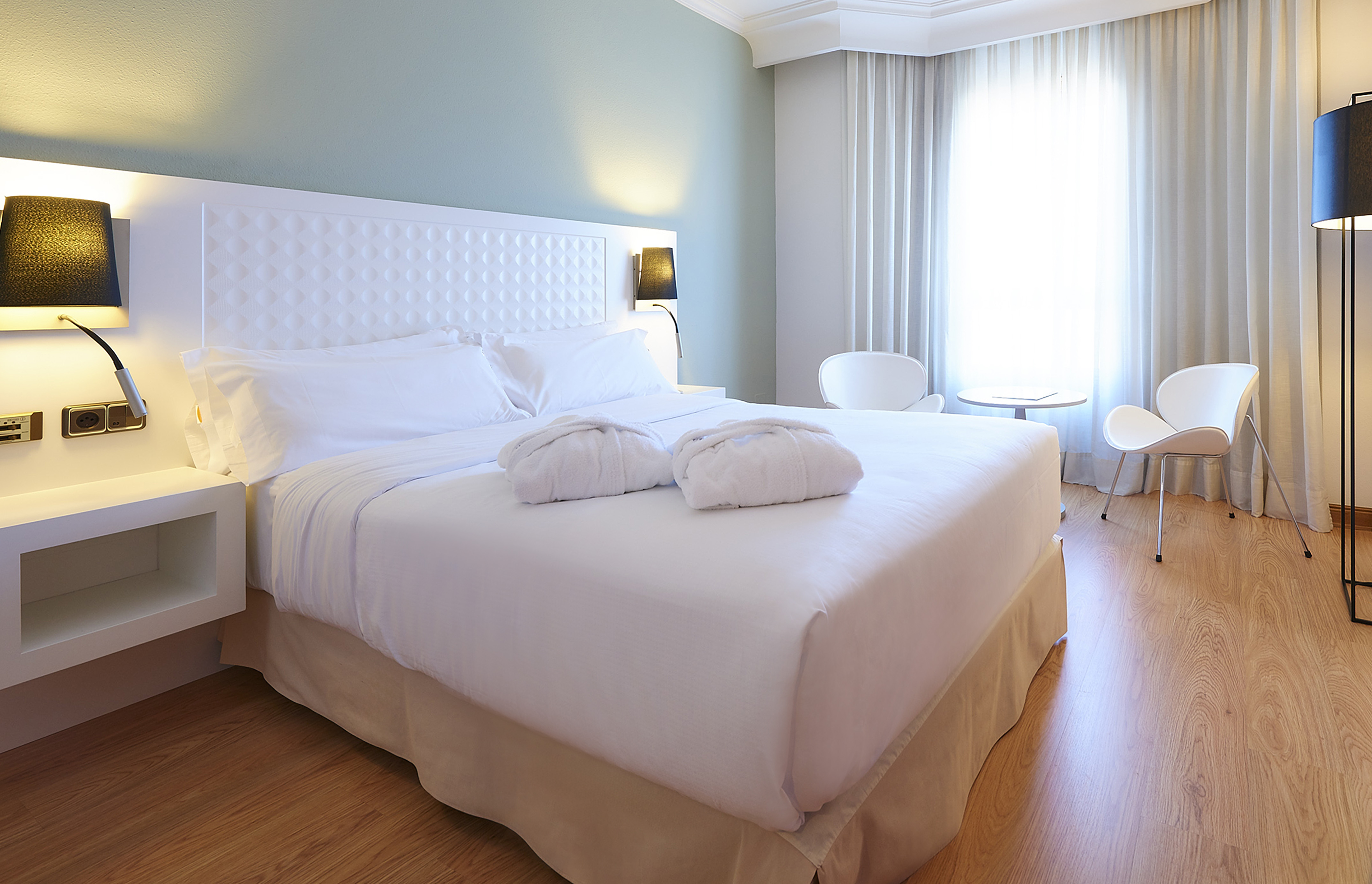 Modern hotel bedroom with white bed, bathrobes, wood floor, bright window.