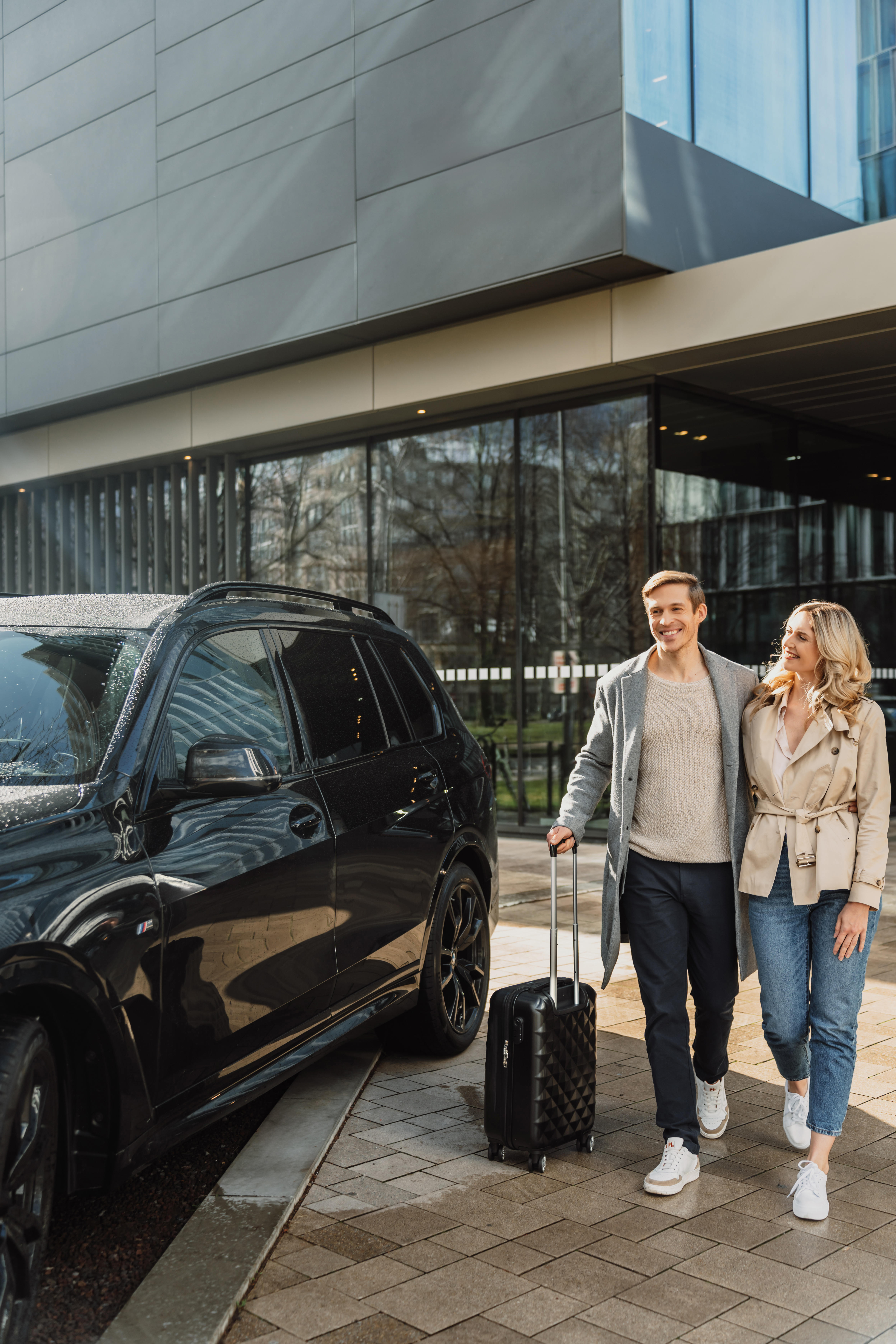 Smiling couple with luggage by luxury black SUV at modern building entrance.