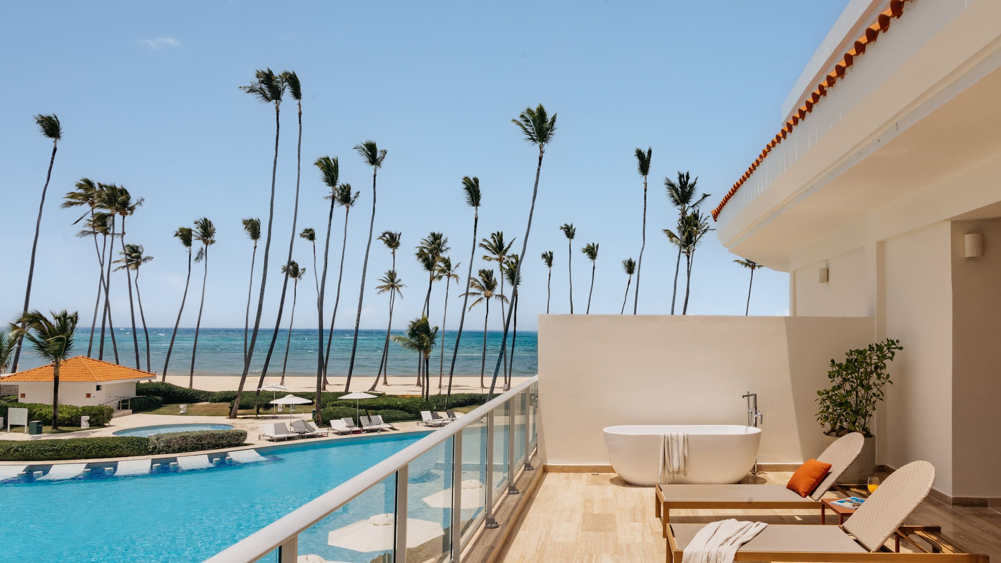 Luxury hotel balcony with private tub, pool, palm trees, and ocean view.