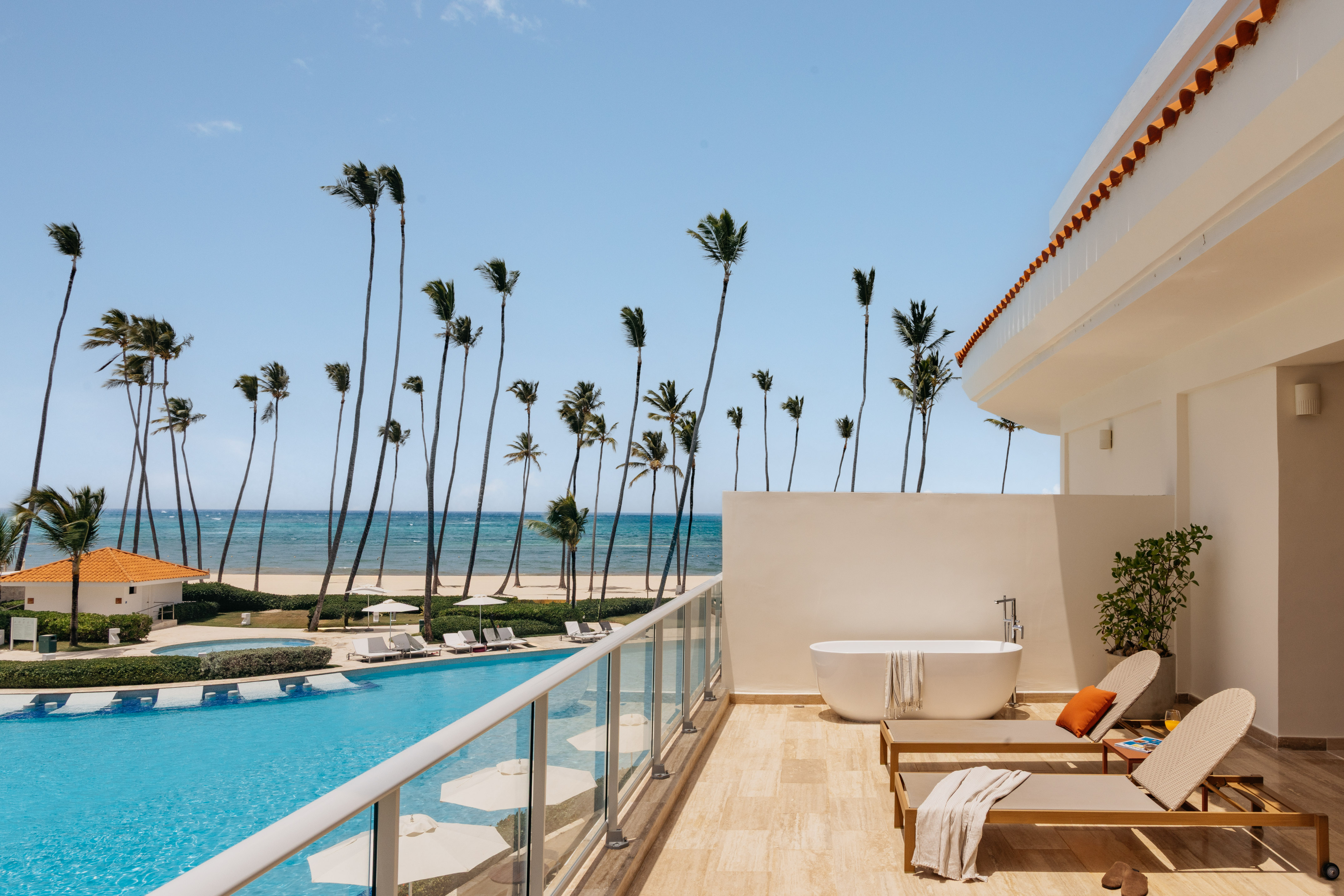 Luxury hotel balcony with private tub, pool, palm trees, and ocean view.