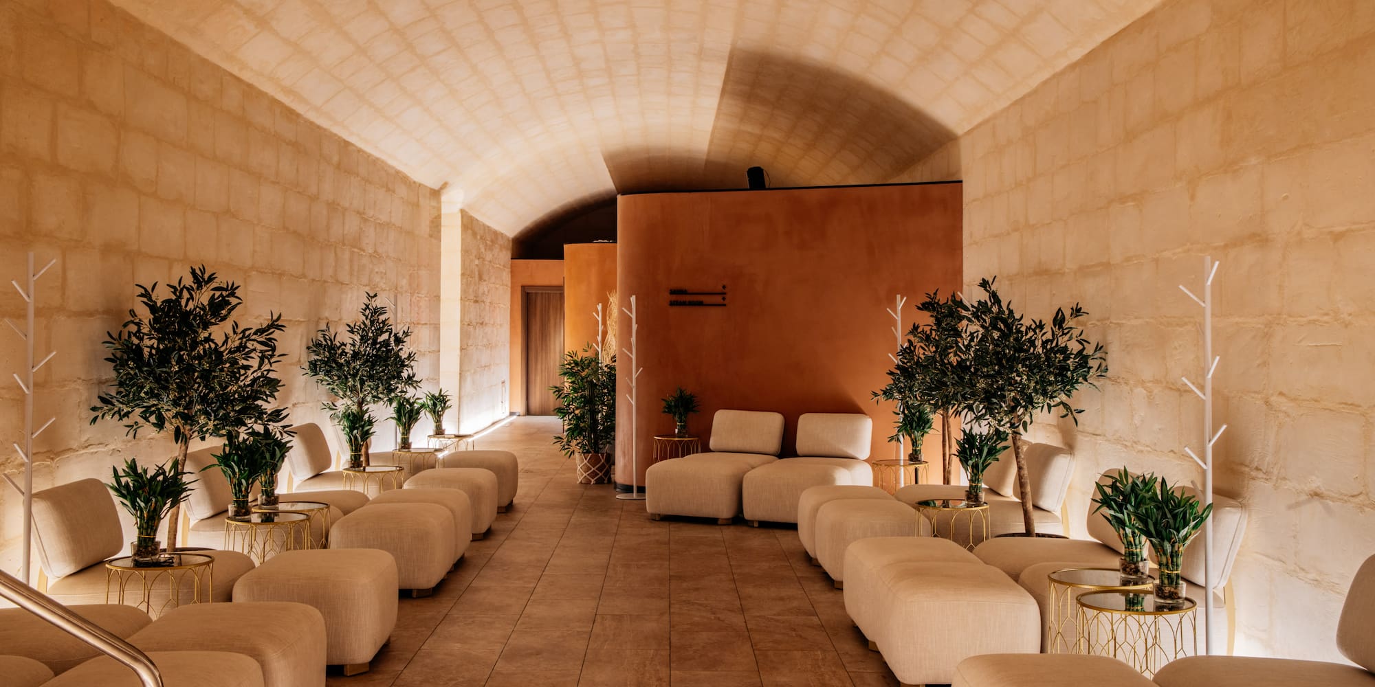 Luxurious spa lounge with vaulted stone ceiling, cream seating, and potted olive trees.