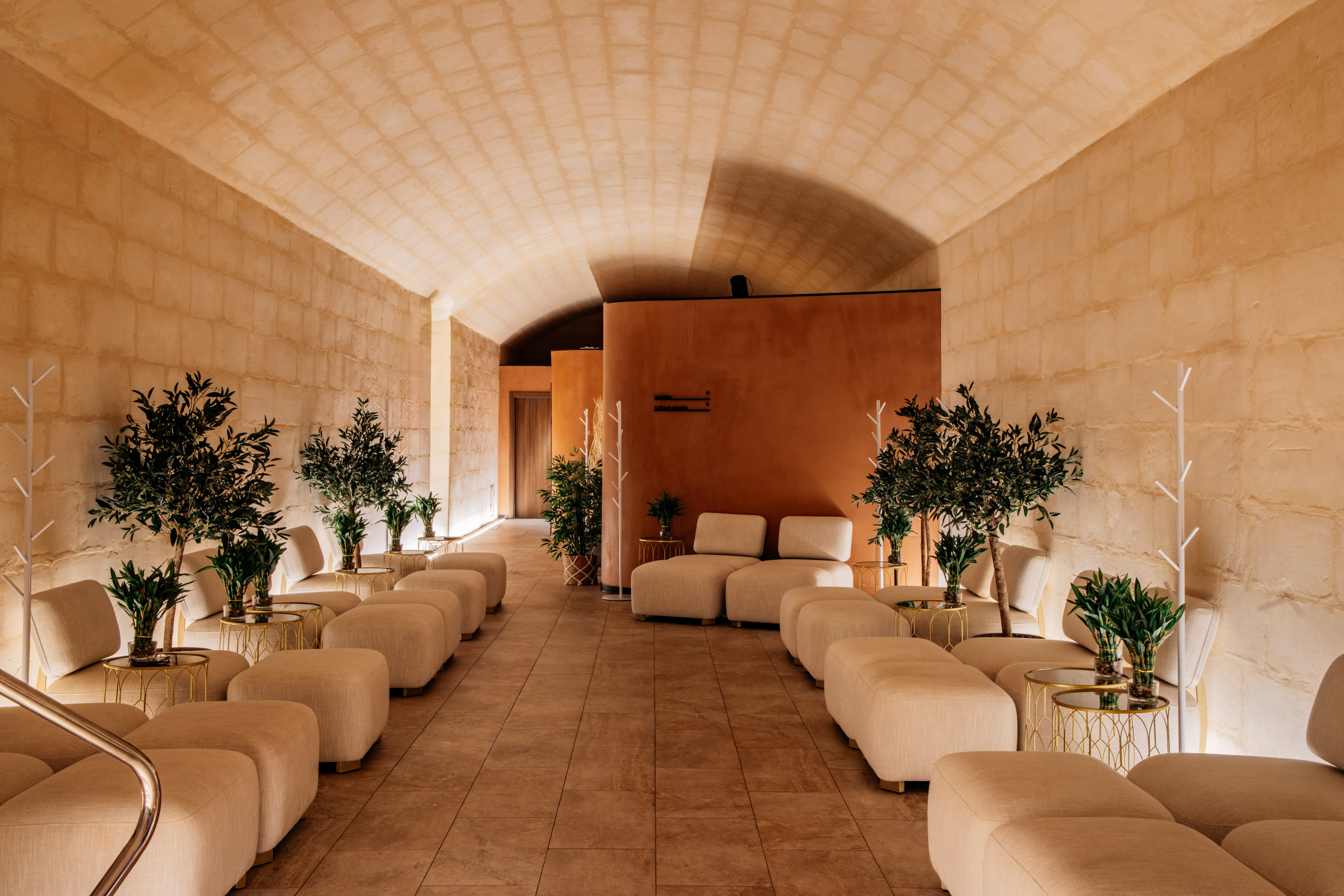 Luxurious spa lounge with vaulted stone ceiling, cream seating, and potted olive trees.