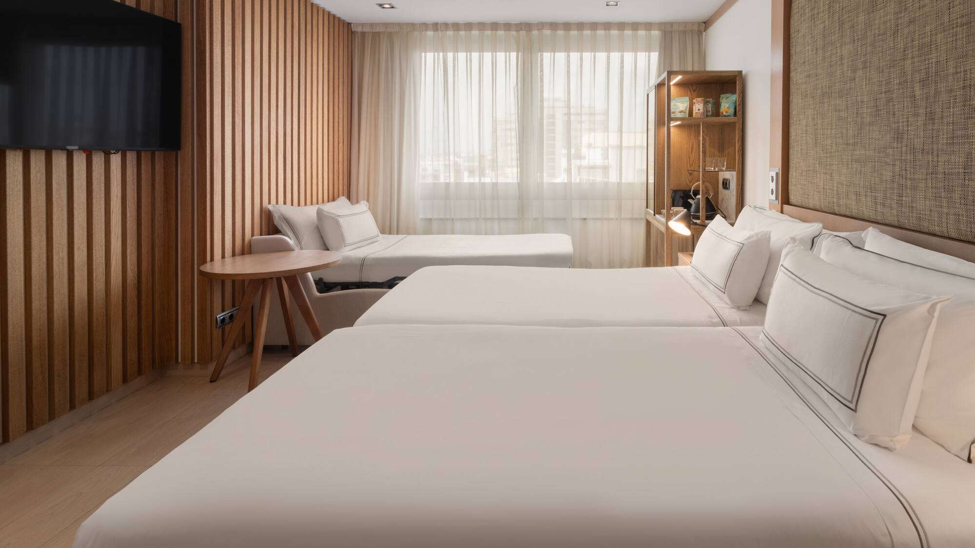 Modern hotel room with twin beds, a sofa bed, wood paneling, and TV.