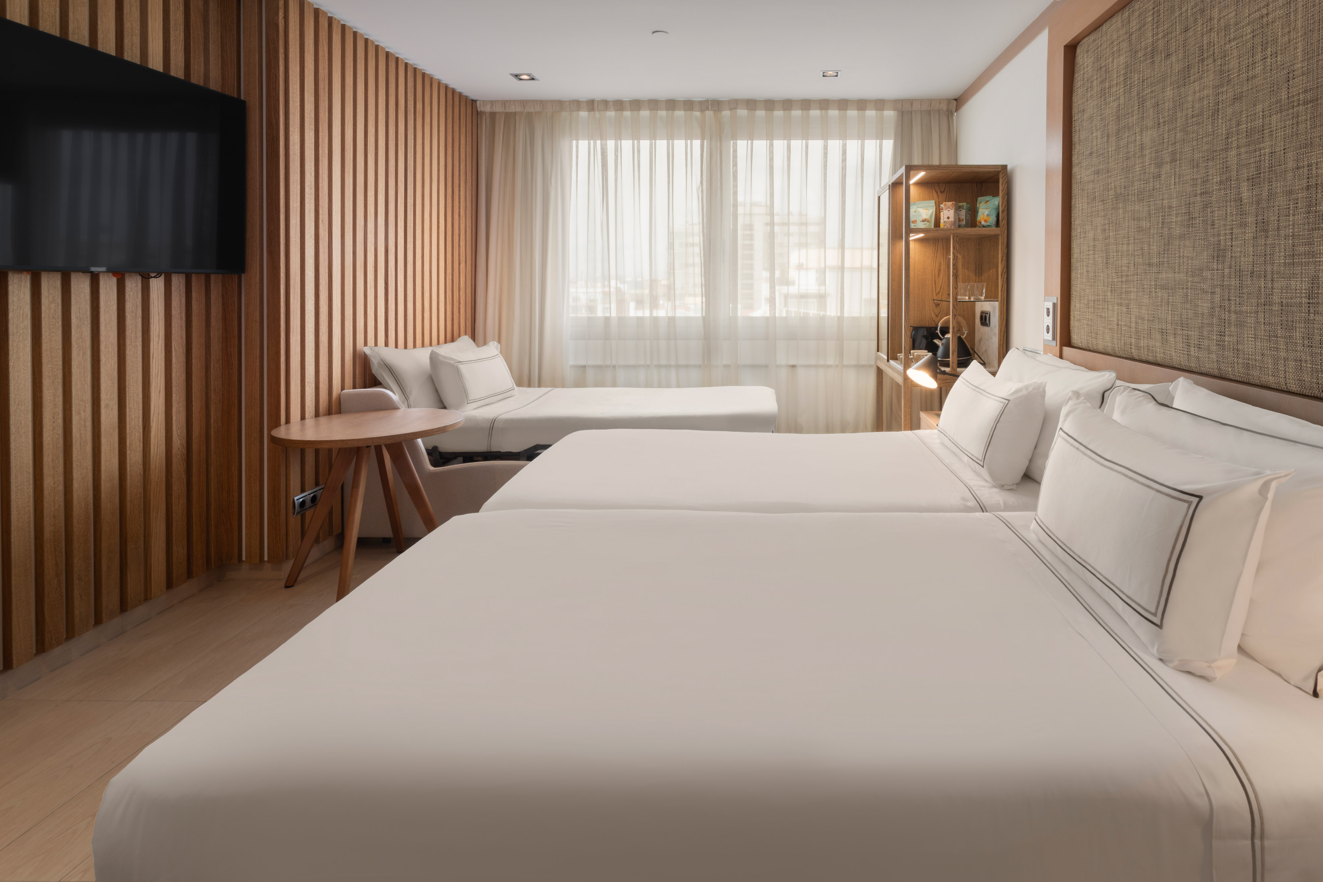 Modern hotel room with twin beds, a sofa bed, wood paneling, and TV.