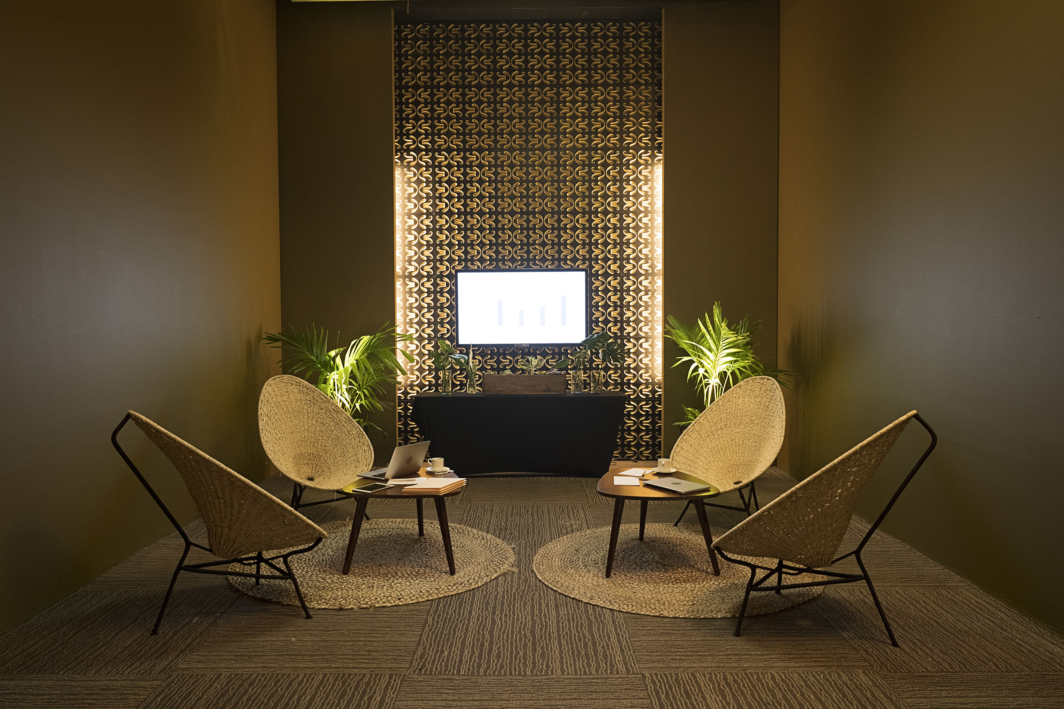 Modern lounge with wicker chairs, tables, plants, and presentation screen.