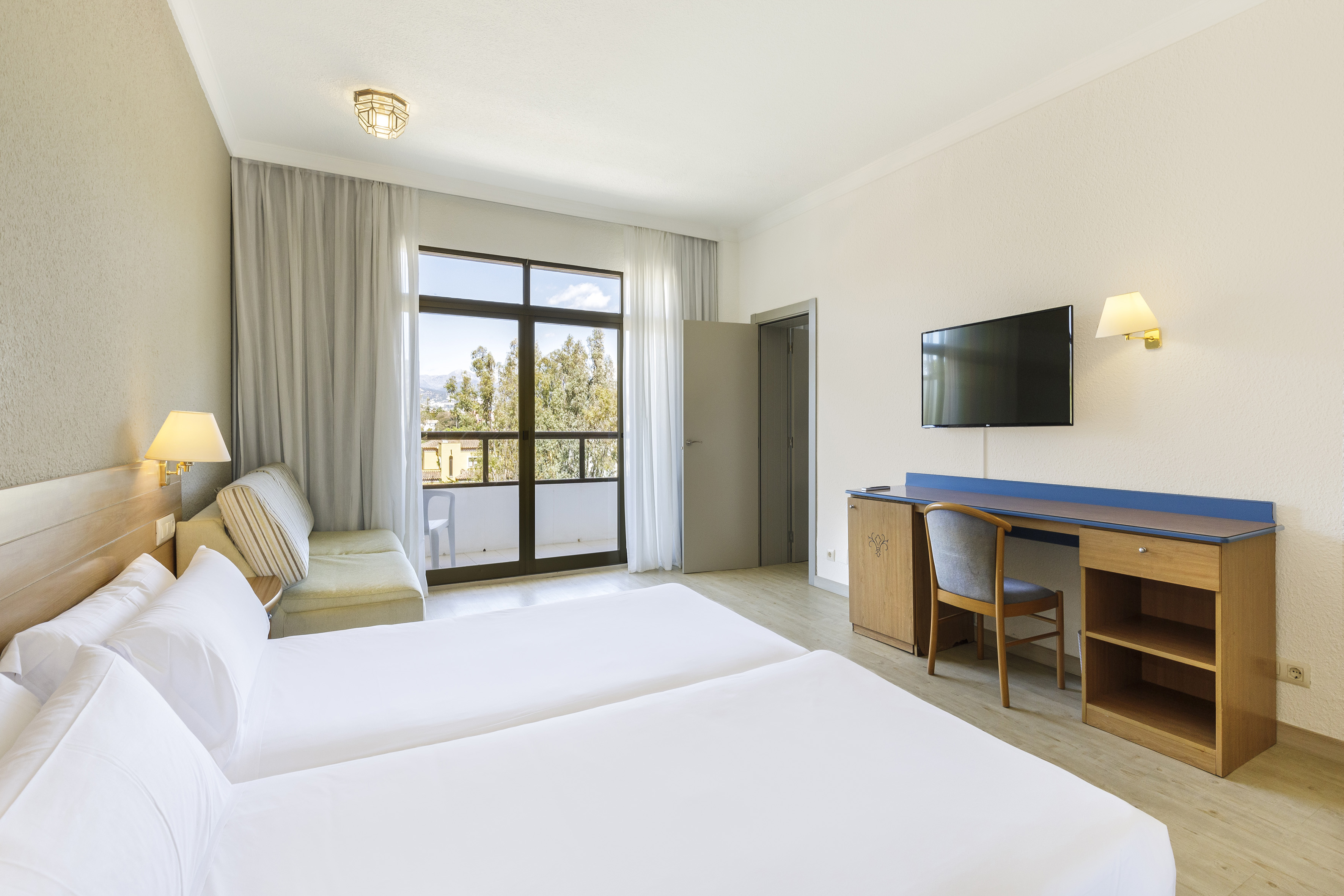 Hotel room with twin beds, sofa, desk, TV, and balcony view of mountains.