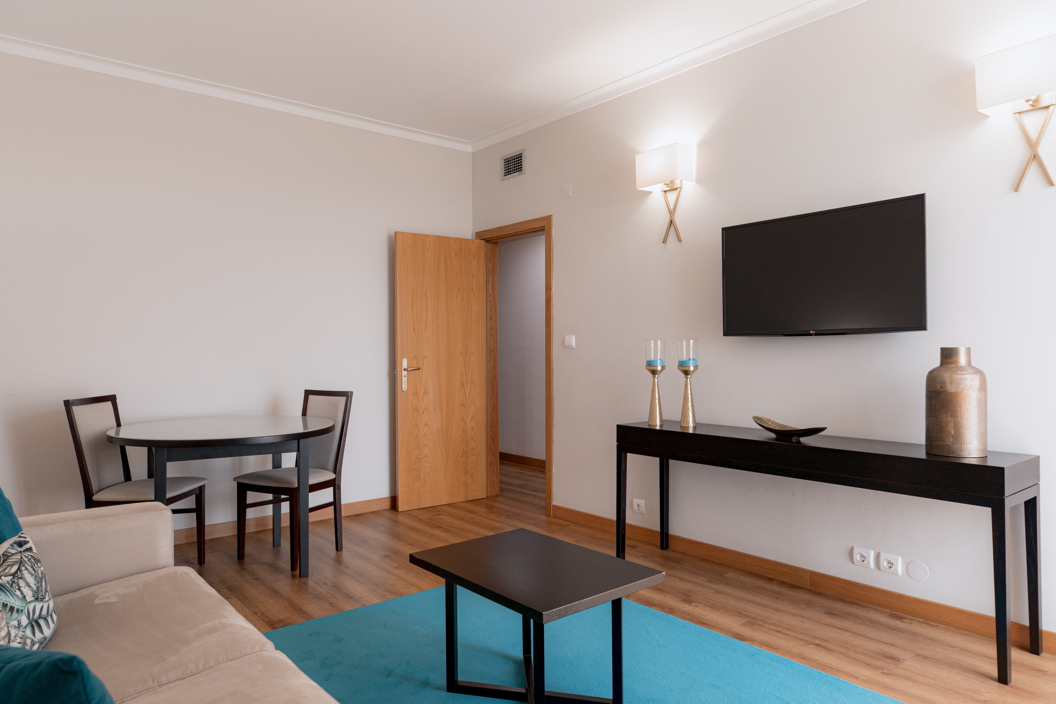 Modern living room: sofa, blue rug, coffee table, dining area, wall TV.