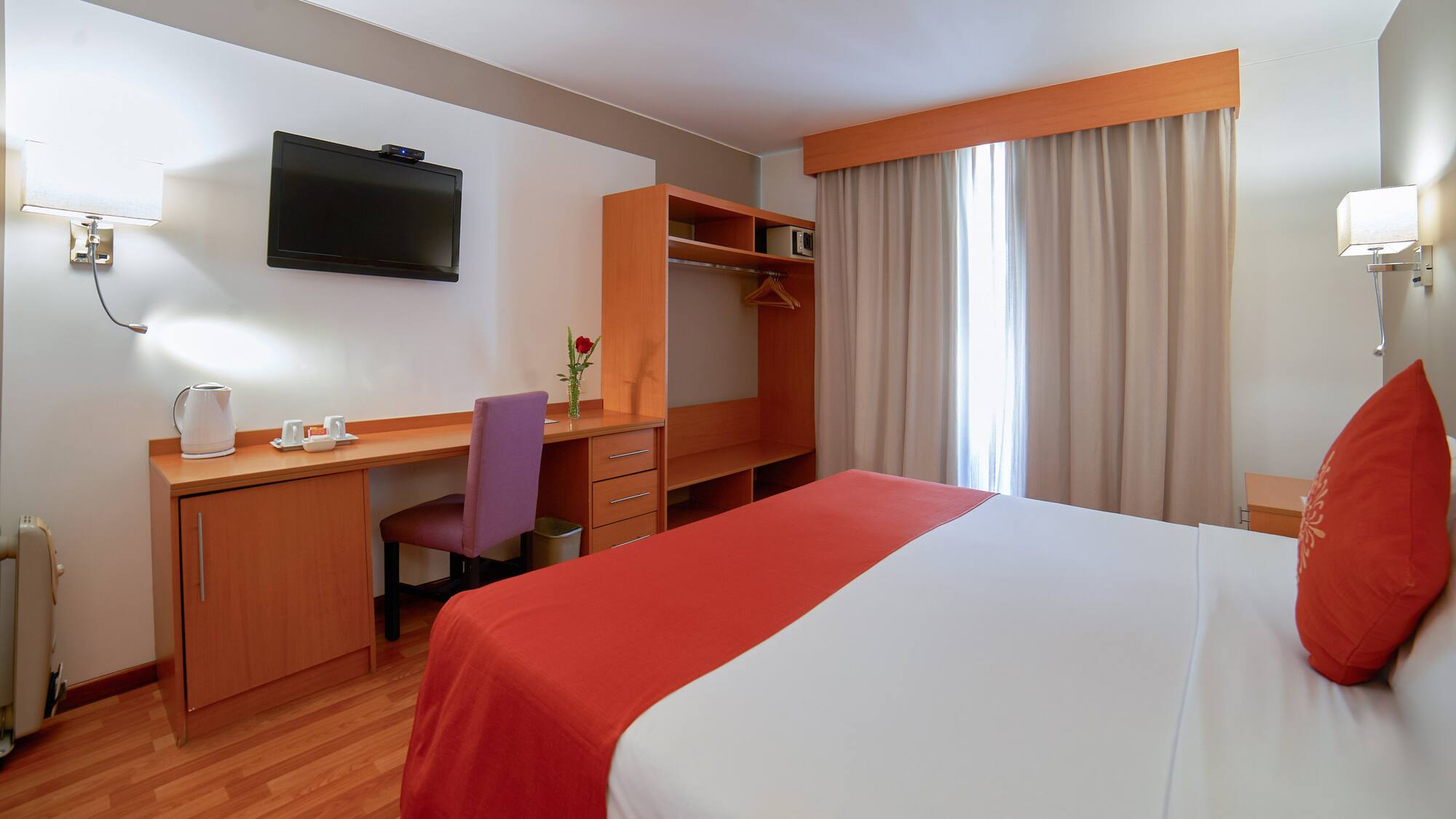 Modern hotel room with bed, desk, TV, and light wood furniture.