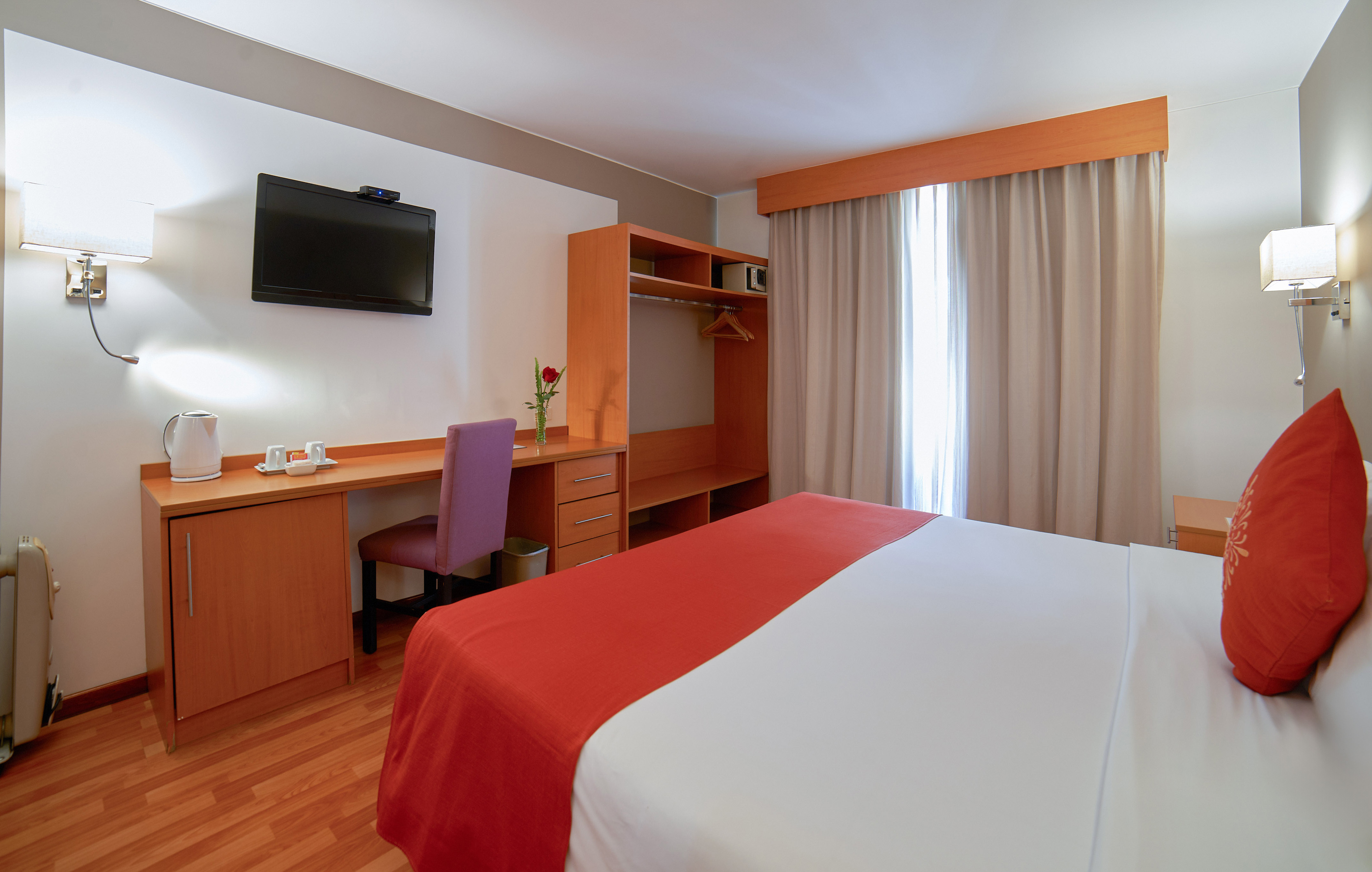 Modern hotel room with bed, desk, TV, and light wood furniture.