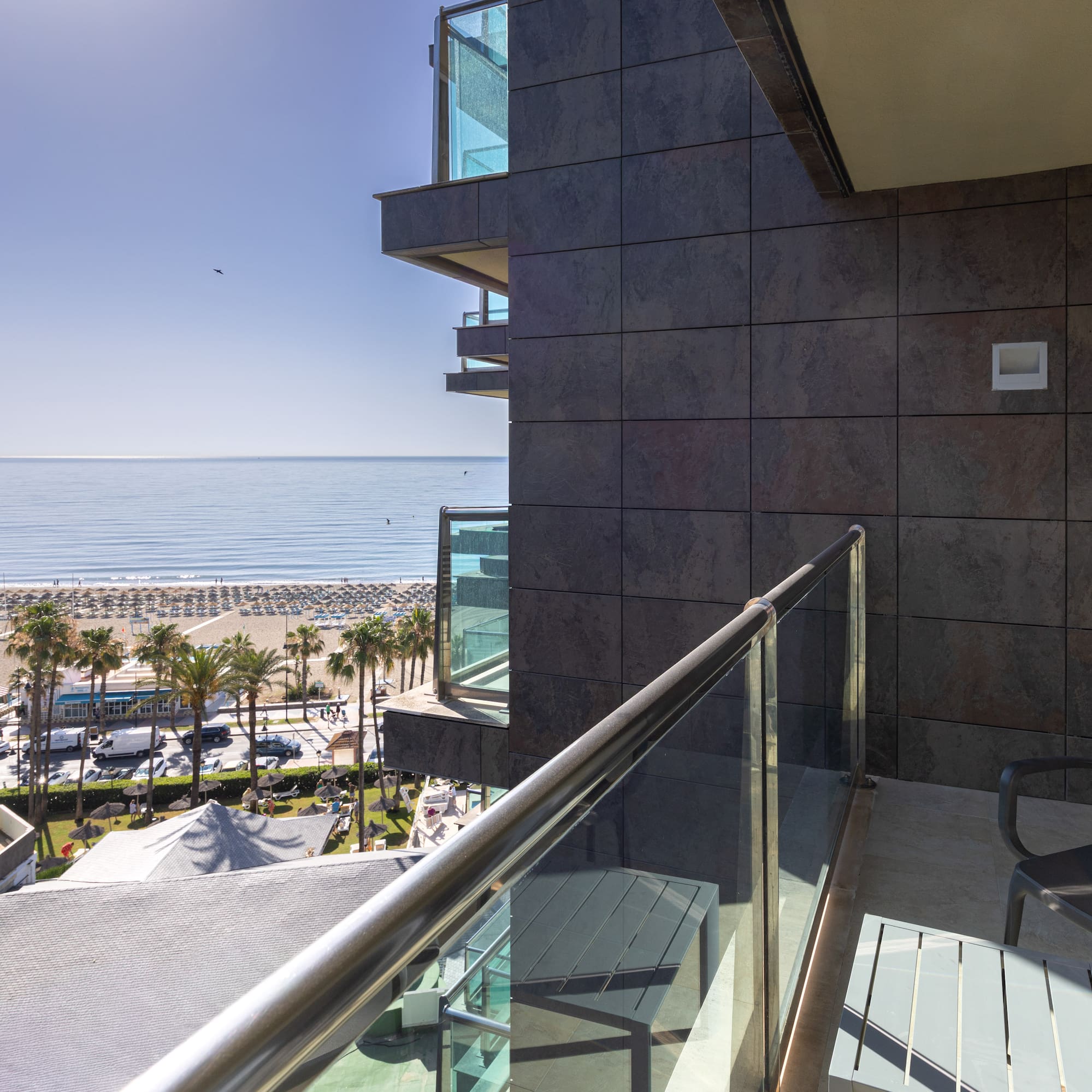 Sunny hotel balcony view of a sandy beach, blue ocean, and palm trees.