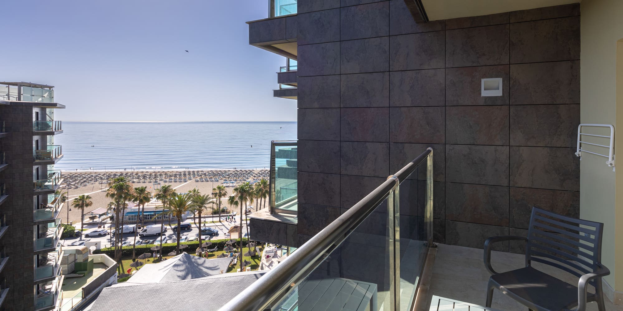 Sunny hotel balcony view of a sandy beach, blue ocean, and palm trees.
