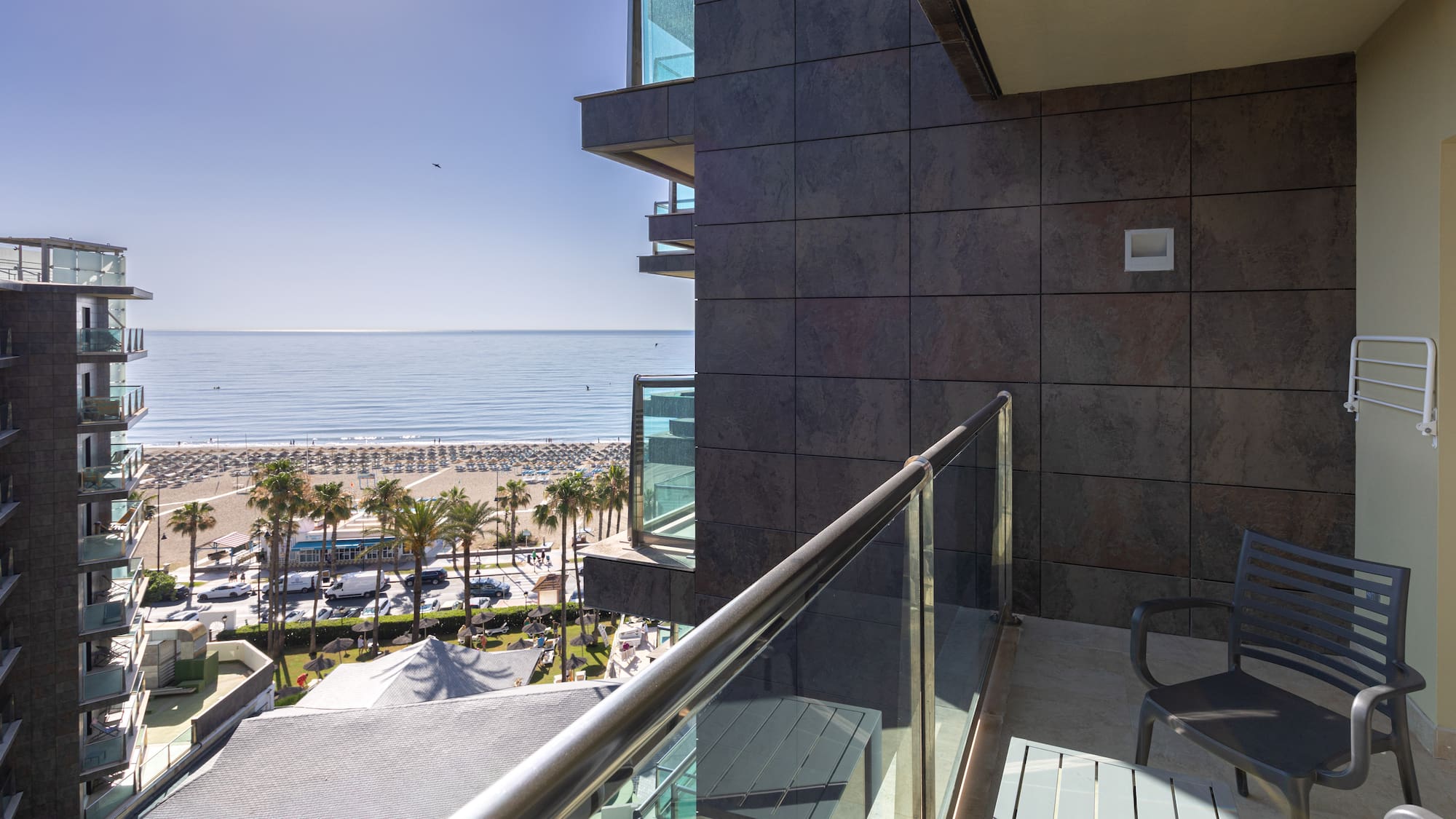 Sunny hotel balcony view of a sandy beach, blue ocean, and palm trees.