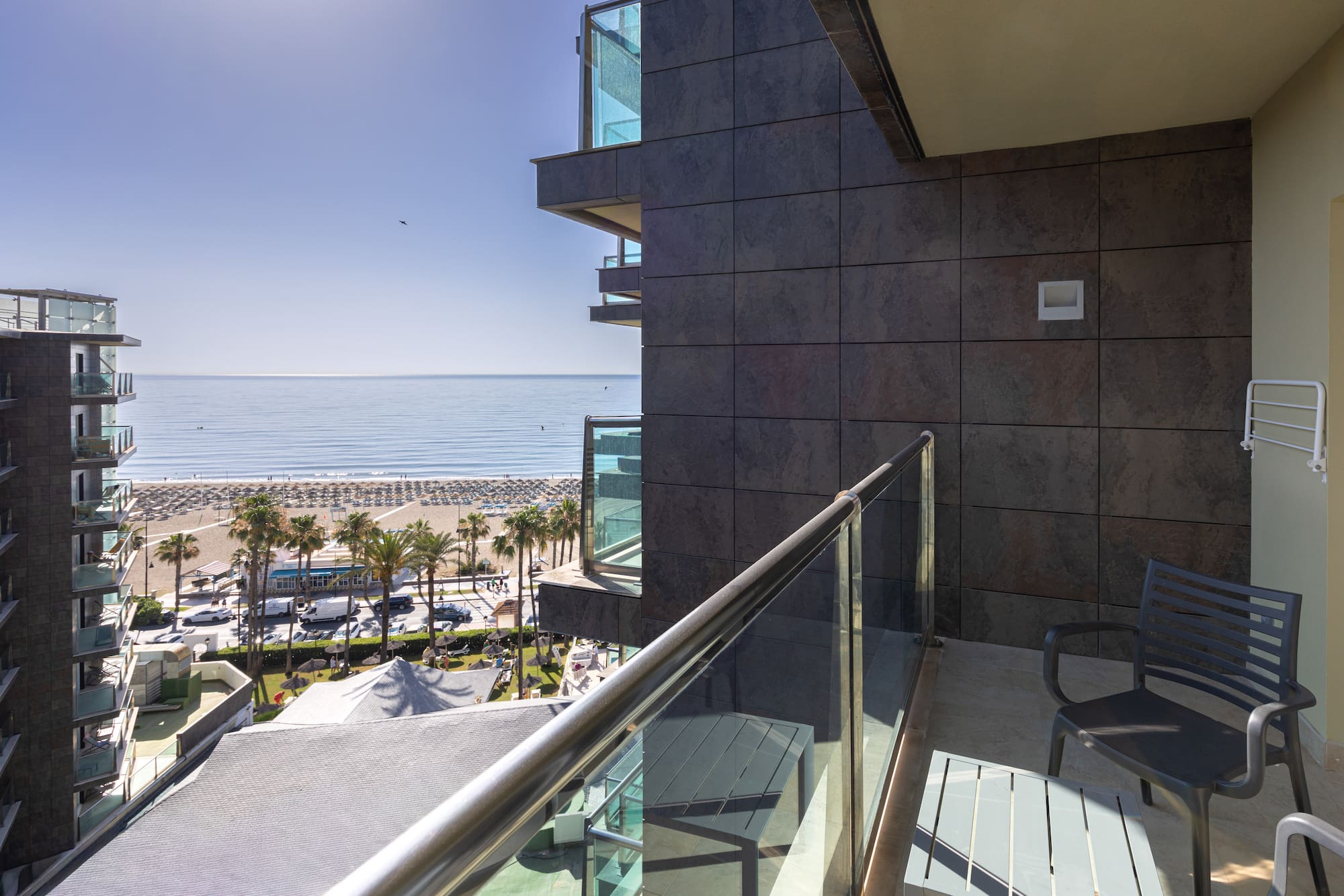 Hotel balcony overlooking a sunny resort beach with palm trees and ocean.